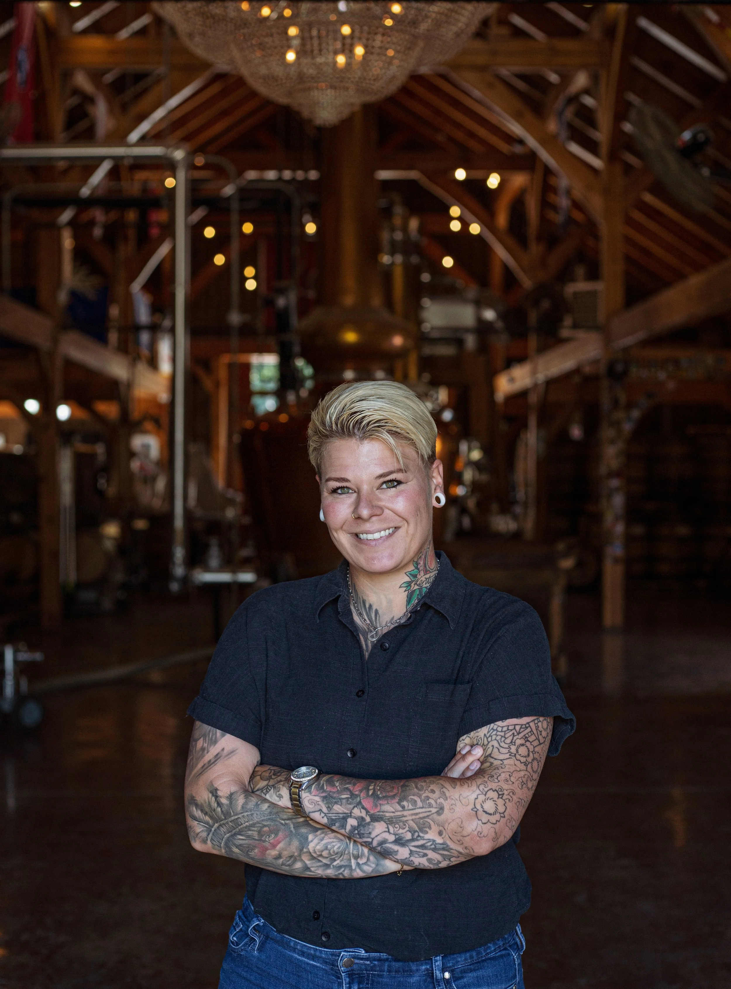 A smiling woman with tattoos, short blonde hair, and piercings, standing with arms crossed inside a rustic wooden building with warm lighting and chandeliers.