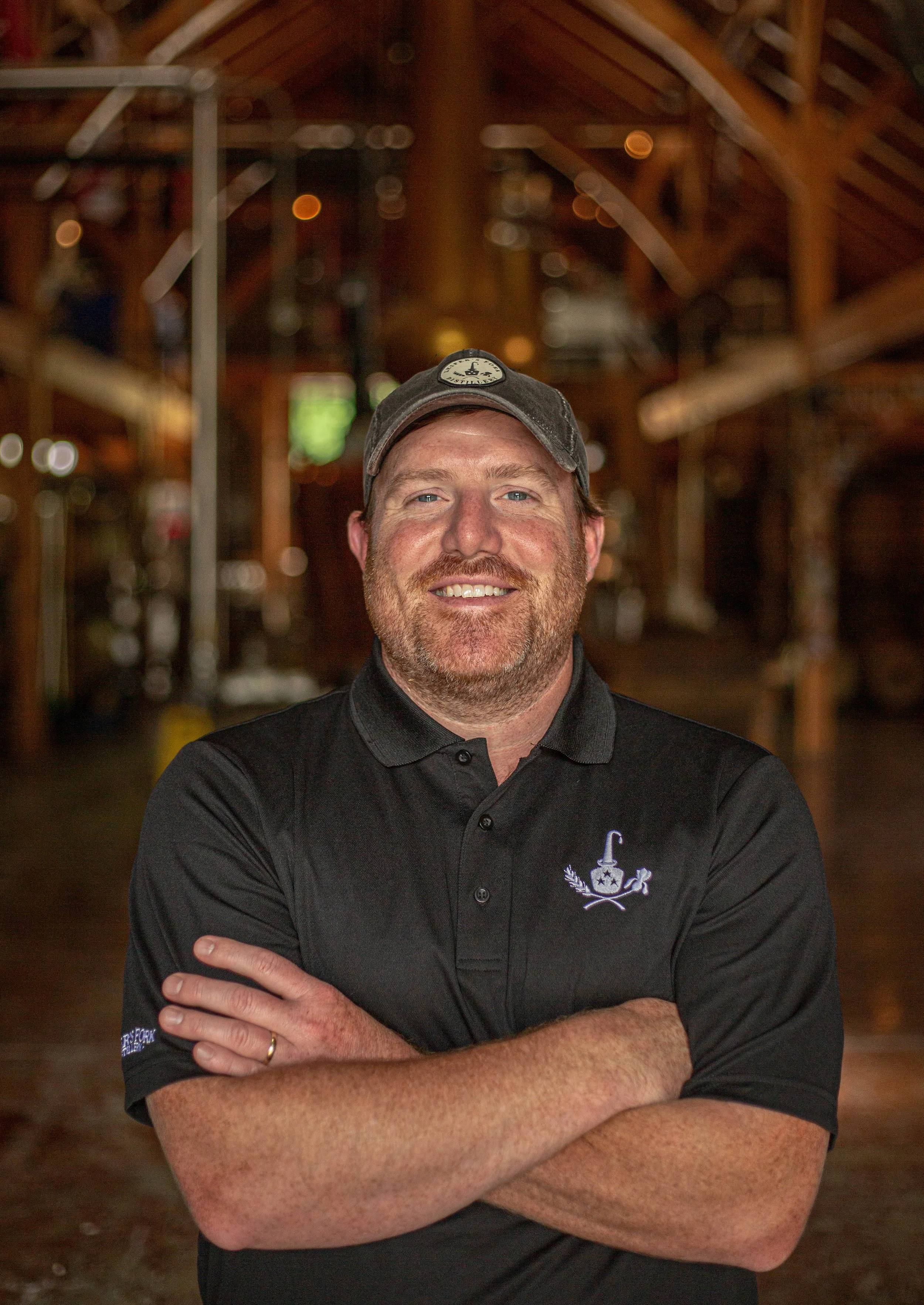 A man with a beard and a baseball cap smiling with arms crossed, standing inside a wooden building or brewery.