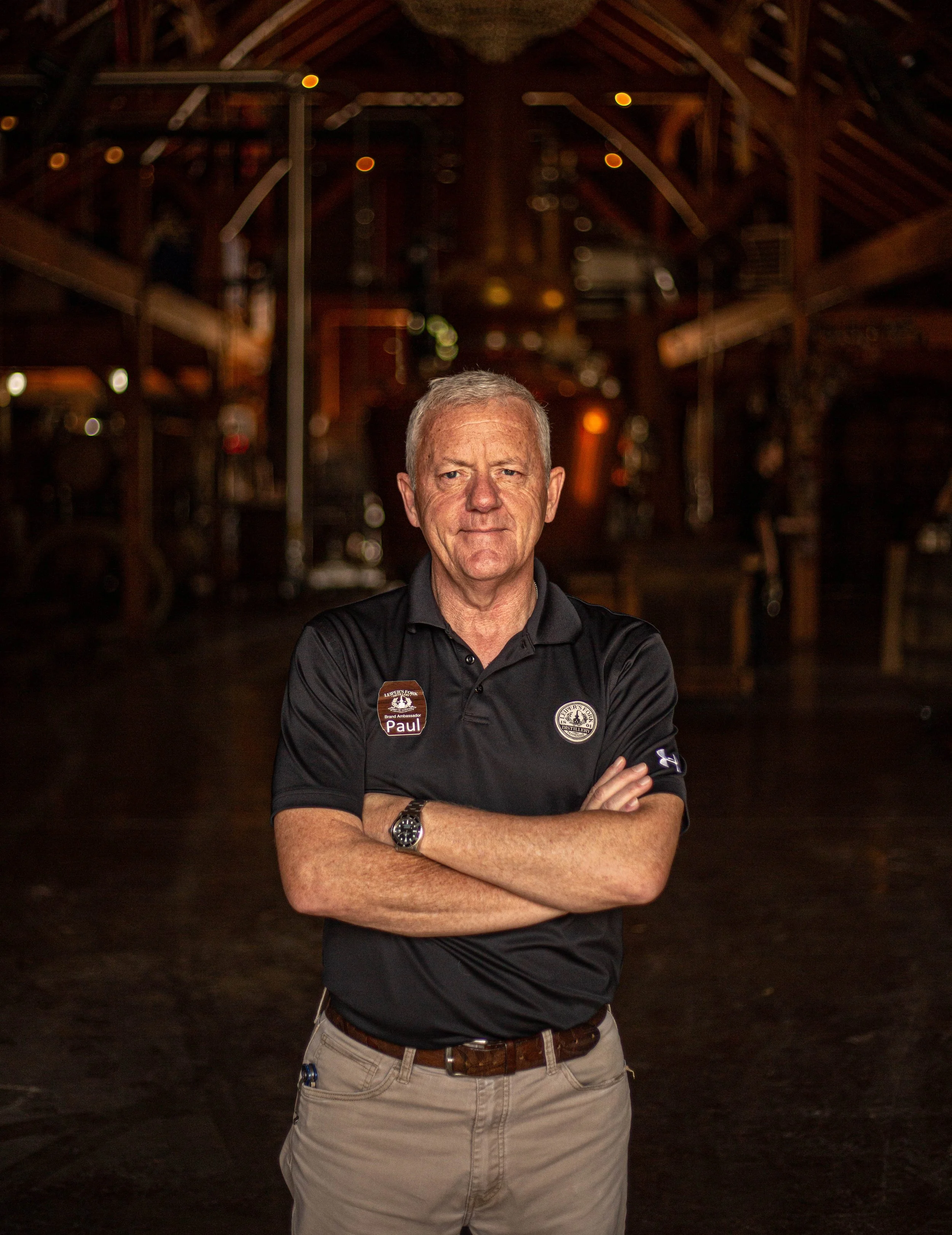 Portrait of an older man with gray hair, standing inside a dimly lit wooden structure with arms crossed, wearing a black polo shirt with logos and khaki pants.