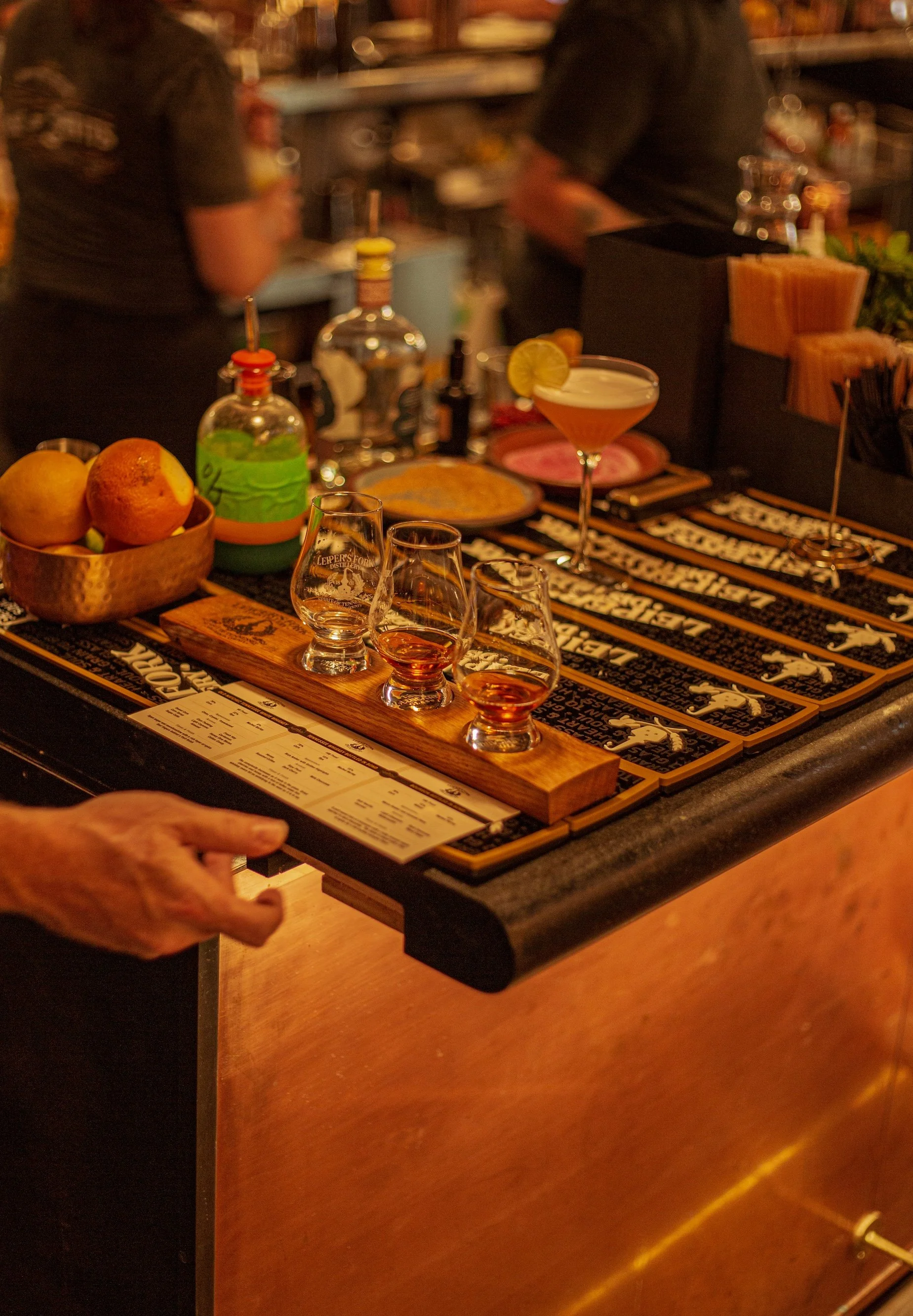 Bar counter with four different types of whiskey, a cocktail with a lemon slice, fresh lemons, and bartending tools, with bartenders in the background.