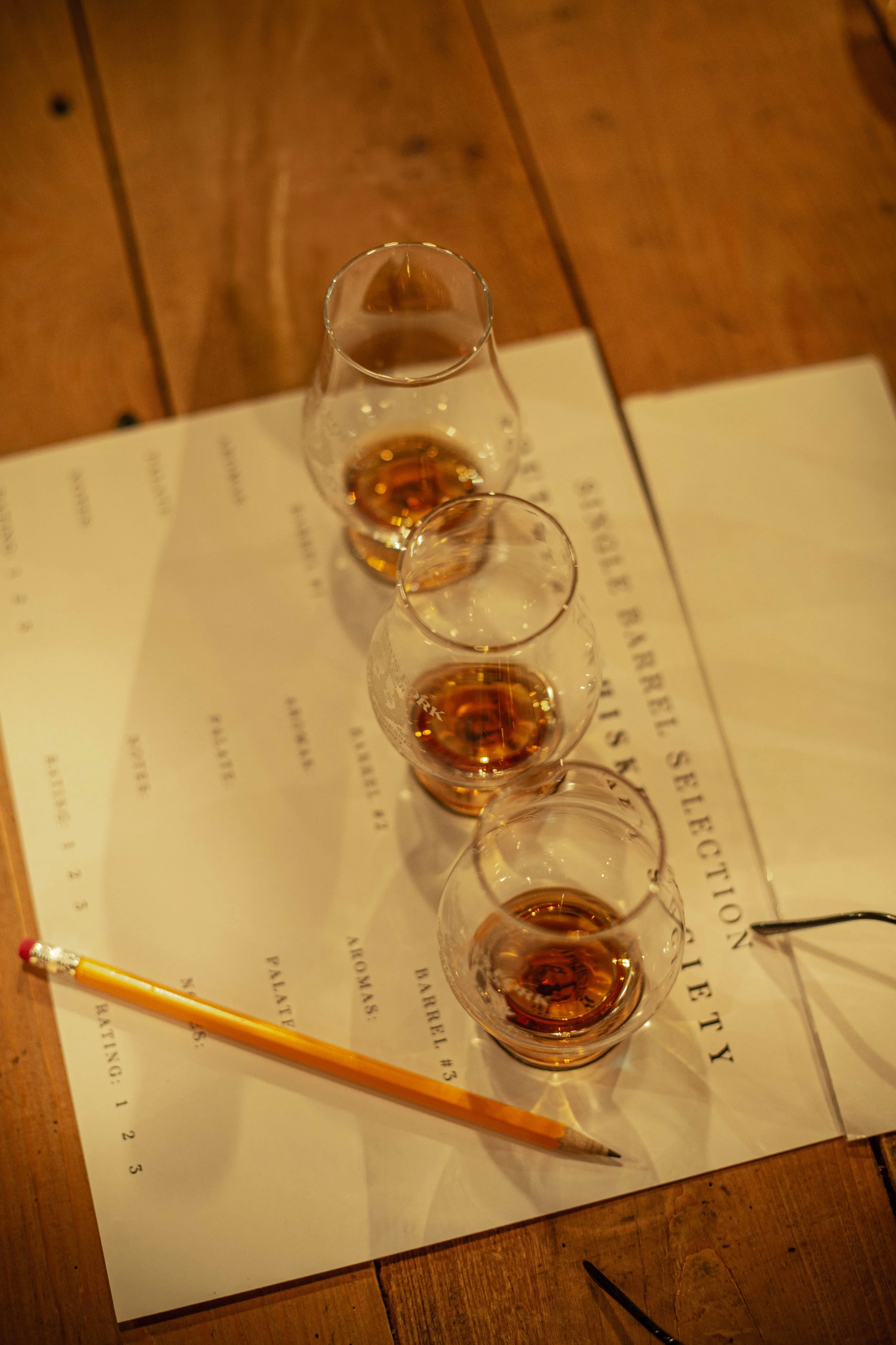Four glasses of amber-colored liquid on a paper with text, brass pencil, and a wooden table.