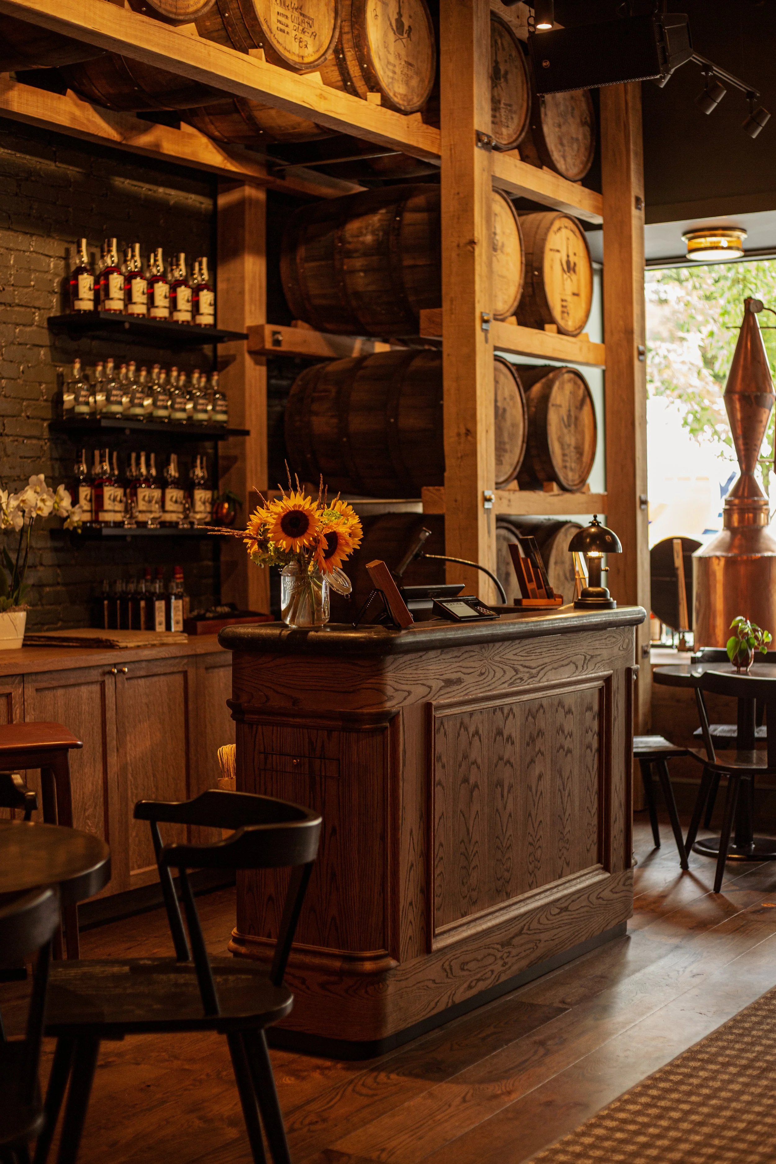 Interior of a rustic bar or restaurant with wooden barrels on shelves, a wooden counter with a sunflower bouquet, and dim lighting.
