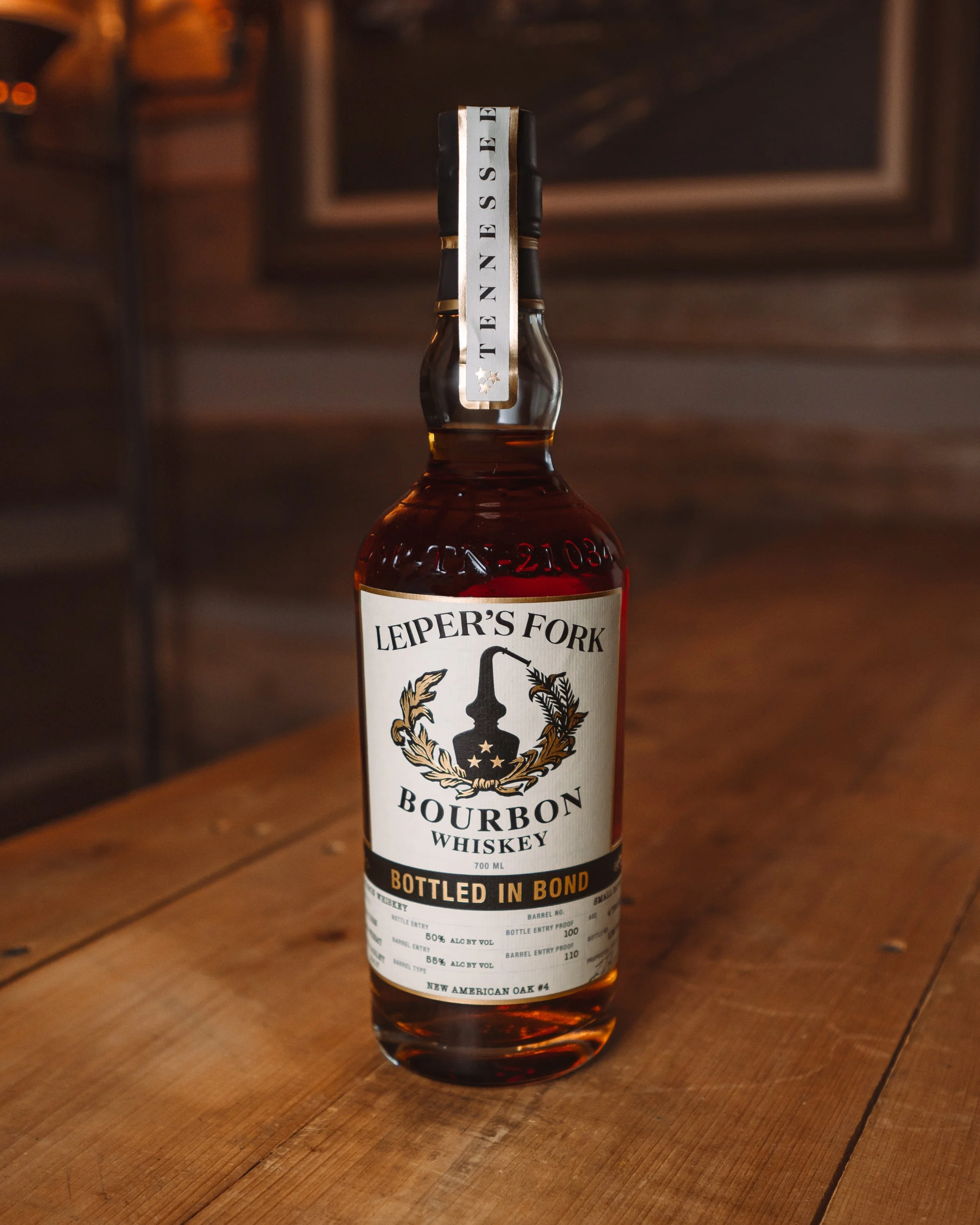 A bottle of Leiper's Fork bourbon whiskey with a bond-tied label on a wooden surface in a dimly lit room.