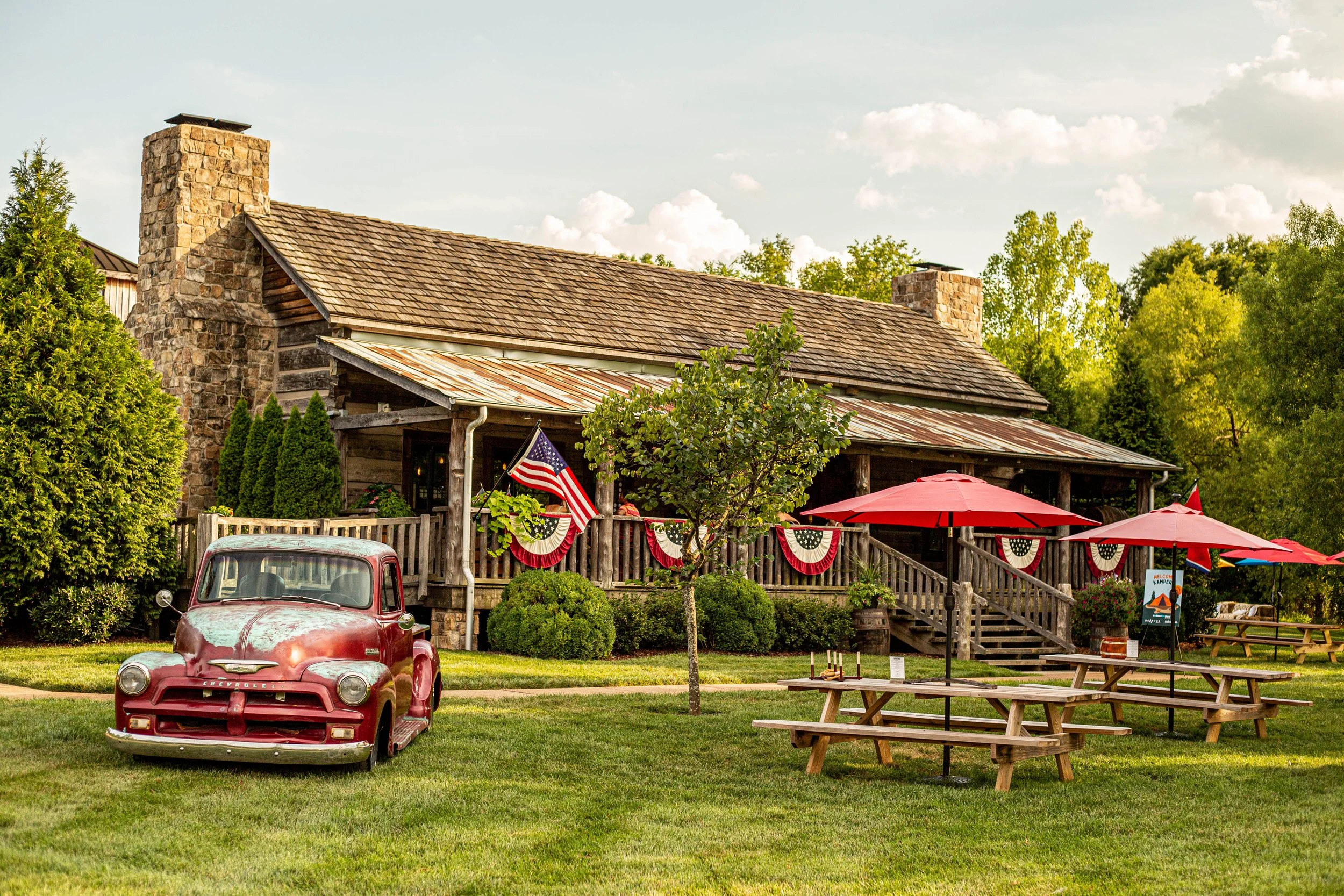 Rustic wooden house decorated with American flags and bunting, with a vintage red Chevrolet in front, outdoor picnic tables with red umbrellas, and lush green trees around, celebrating a patriotic or festive occasion.