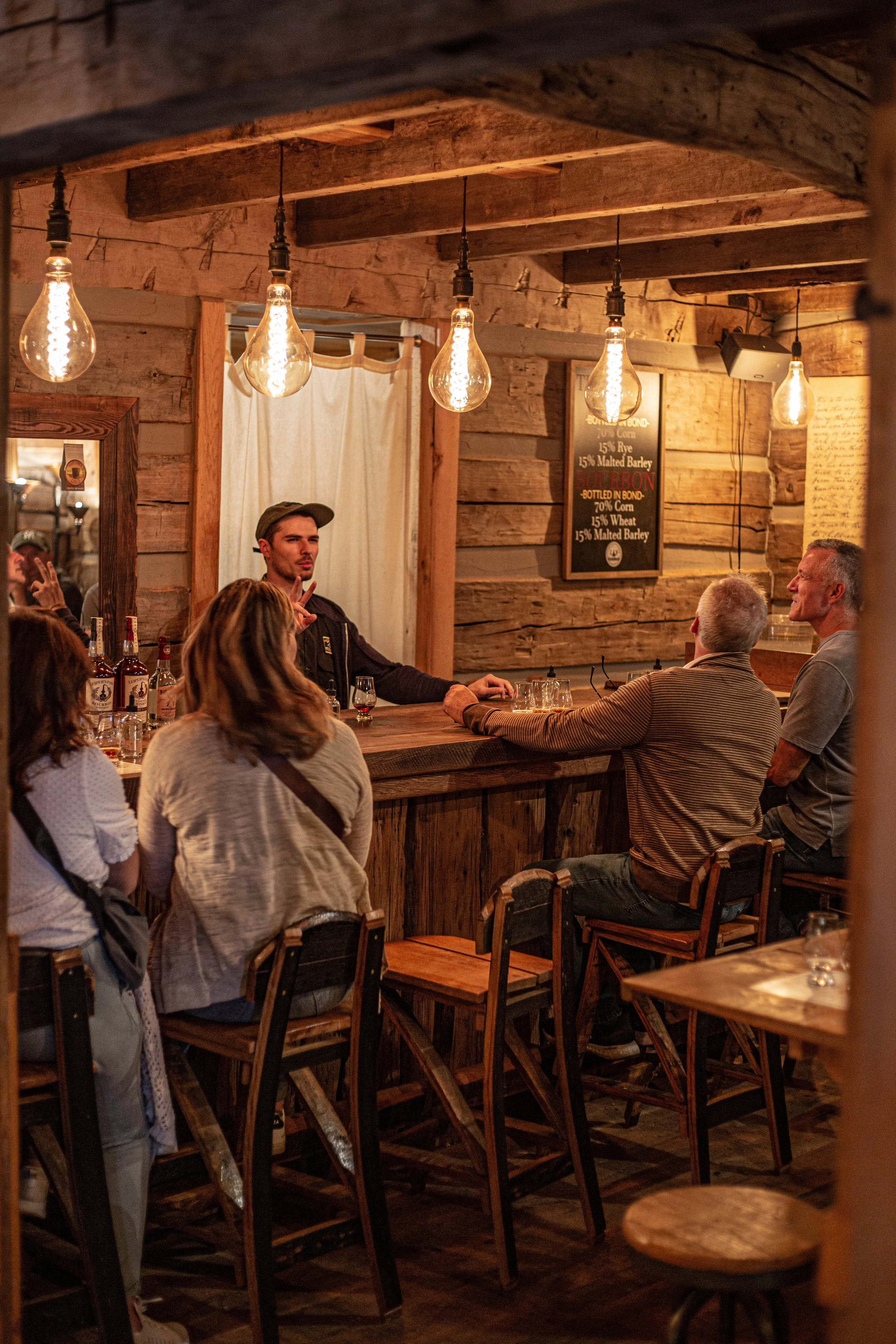 People sitting at a bar in a rustic wooden restaurant, with hanging Edison-style light bulbs overhead, having drinks and conversation.