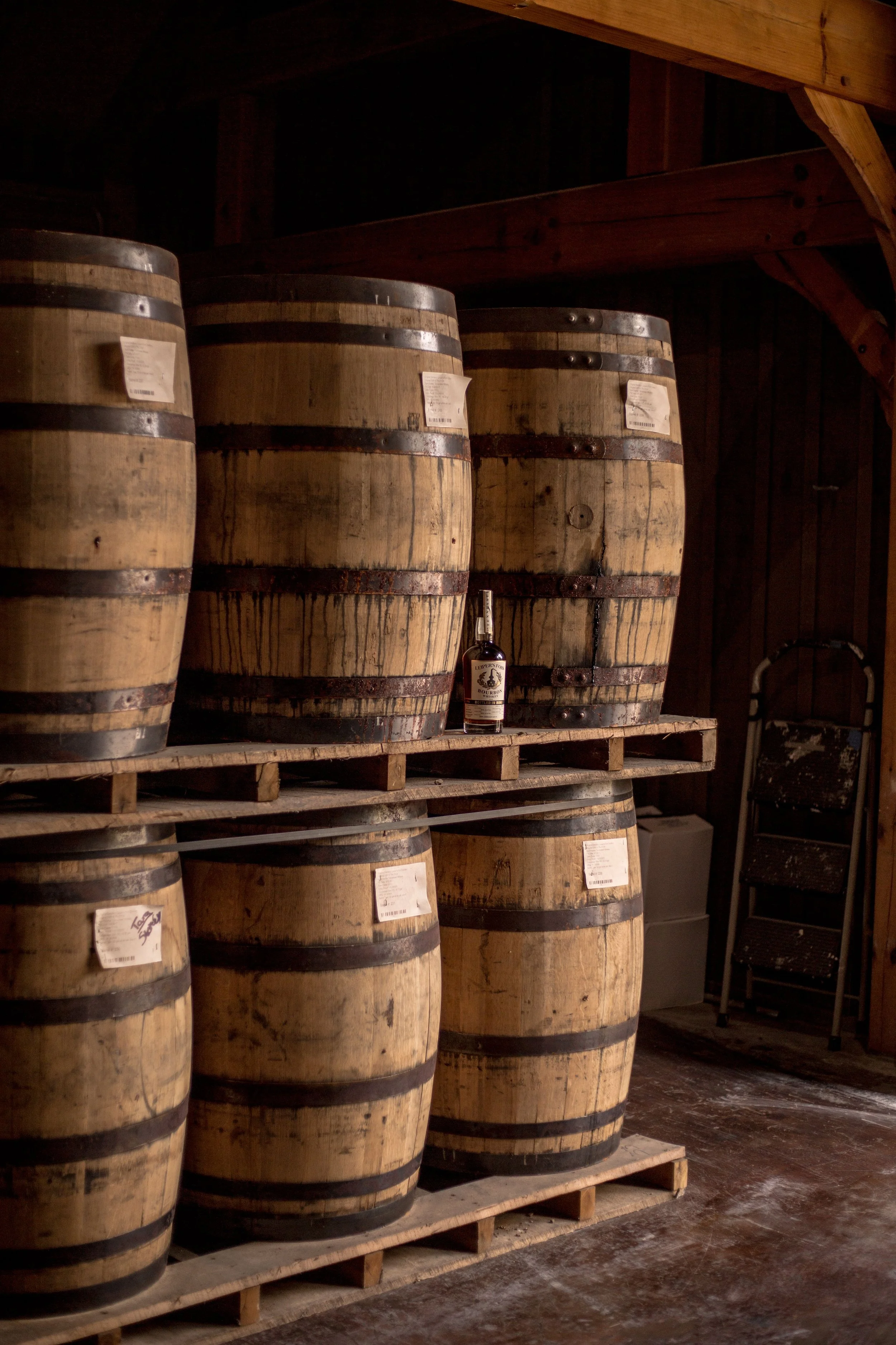 Several large wooden wine barrels on a wooden pallet shelf in a cellar, with a bottle of liquor placed on the shelf among the barrels.