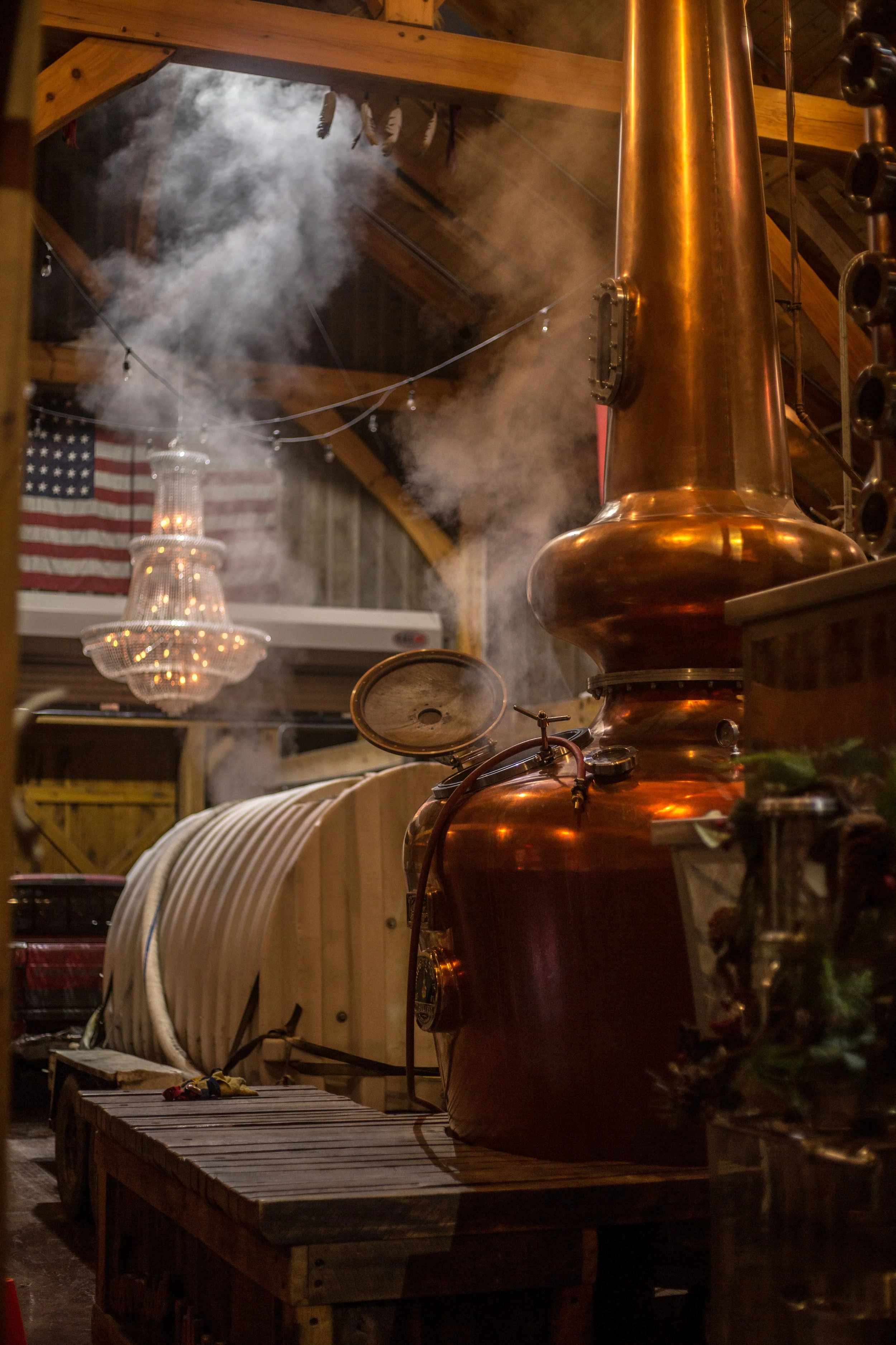 Copper still with steam rising inside a distillery or brewery, American flag hanging in the background, chandelier, wooden walls and ceiling, distillation equipment.