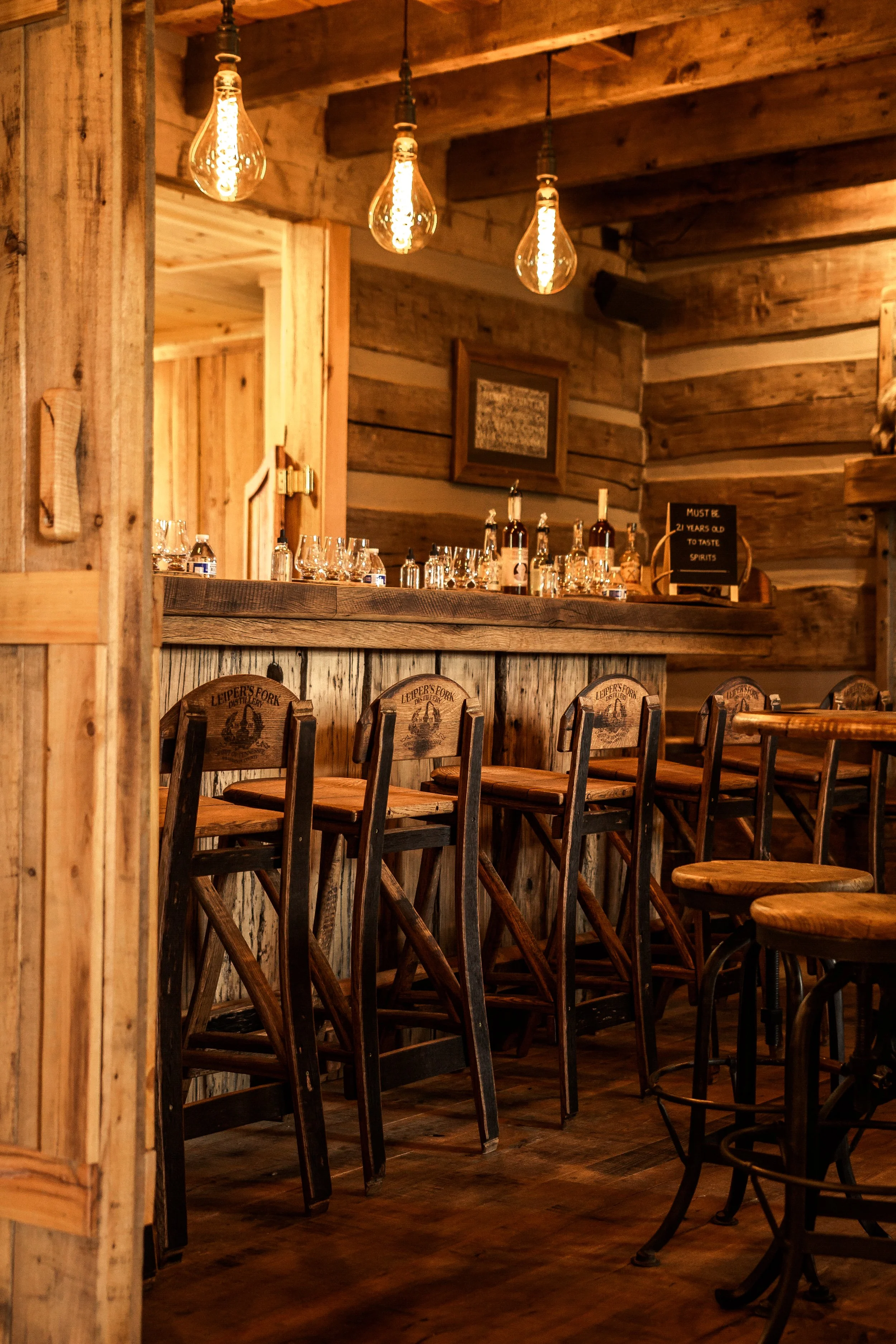 A cozy bar area with wooden furniture and warm lighting, featuring hanging Edison bulbs, a wooden bar counter with bottles and glasses, and rustic wood-paneled walls.