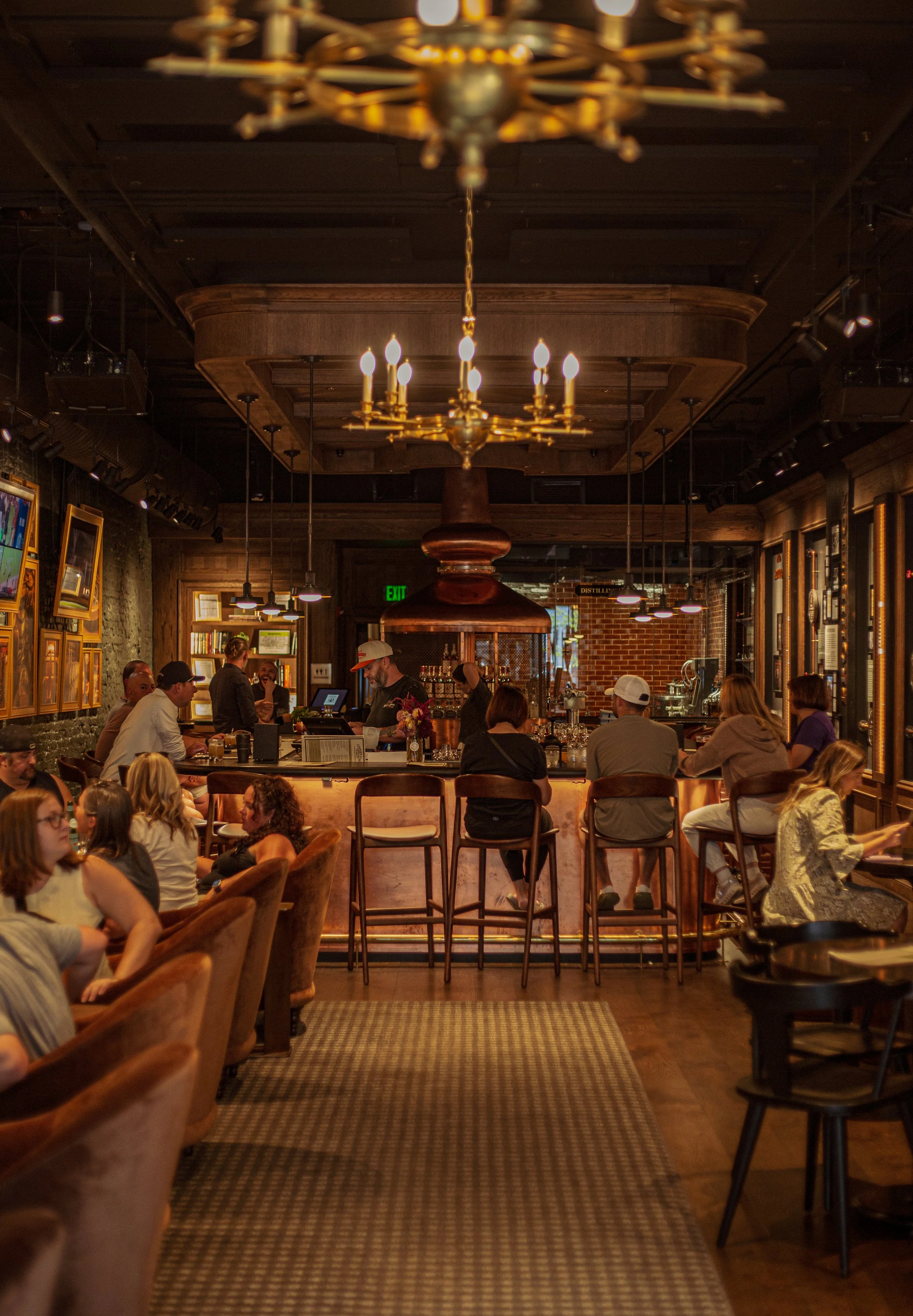 Interior of a cozy bar with warm lighting, wooden decor, and patrons sitting at the bar and tables, some engaging in conversation.