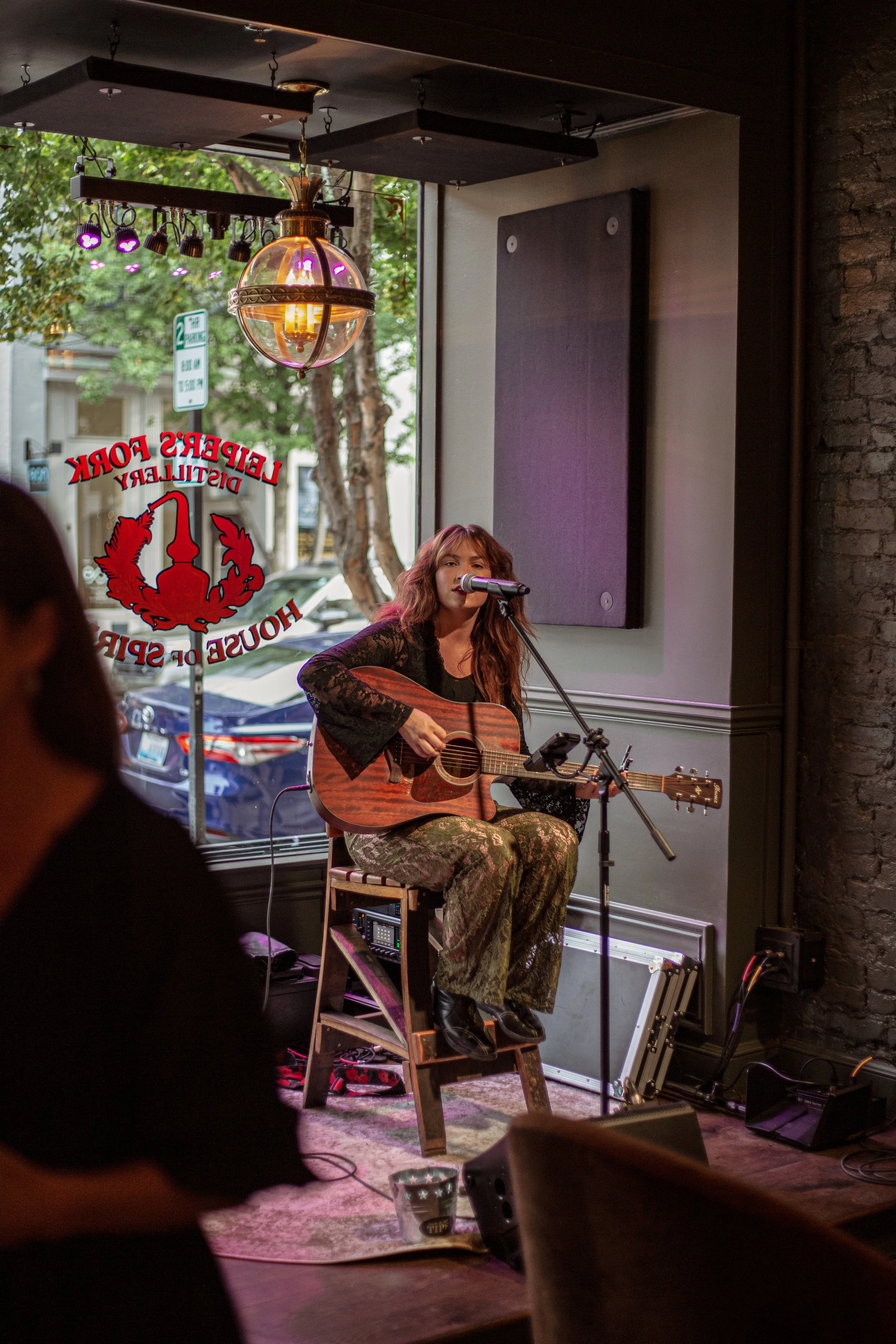 A woman sitting on a stool playing an acoustic guitar and singing into a microphone in a dimly lit cafe. The cafe has a large window with a sign that reads 'Lieber's Fok' and some cars parked outside. There are hanging lights and musical equipment in the background.