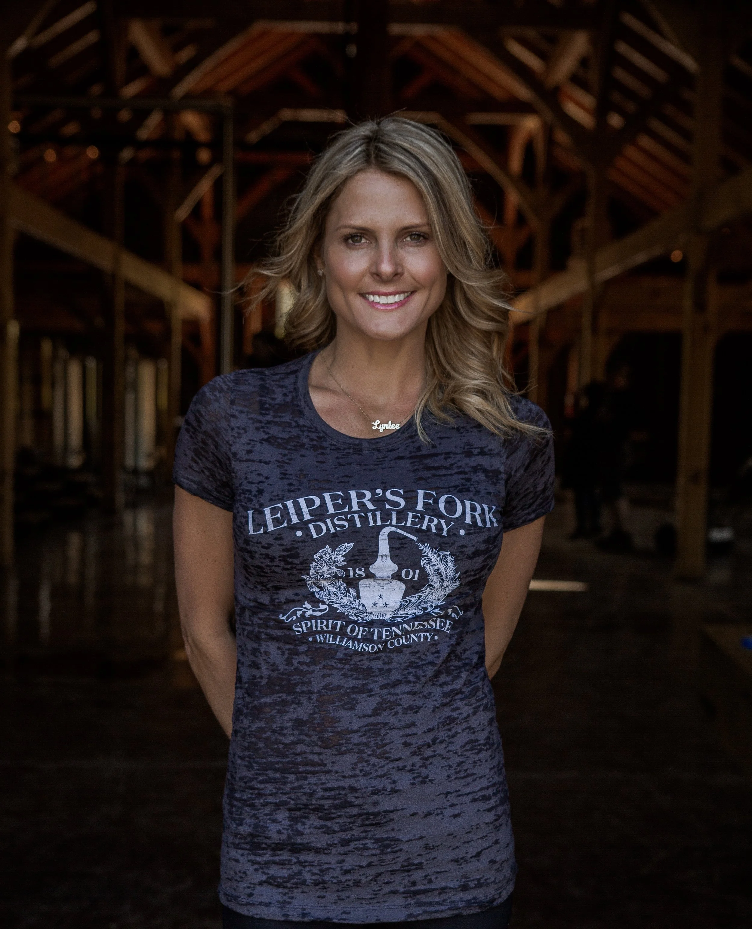 A woman with blonde hair smiling, standing inside a wooden barn, wearing a dark blue T-shirt with 'Leiper's Fork Distillery' logo and a necklace that says 'Lynee'.