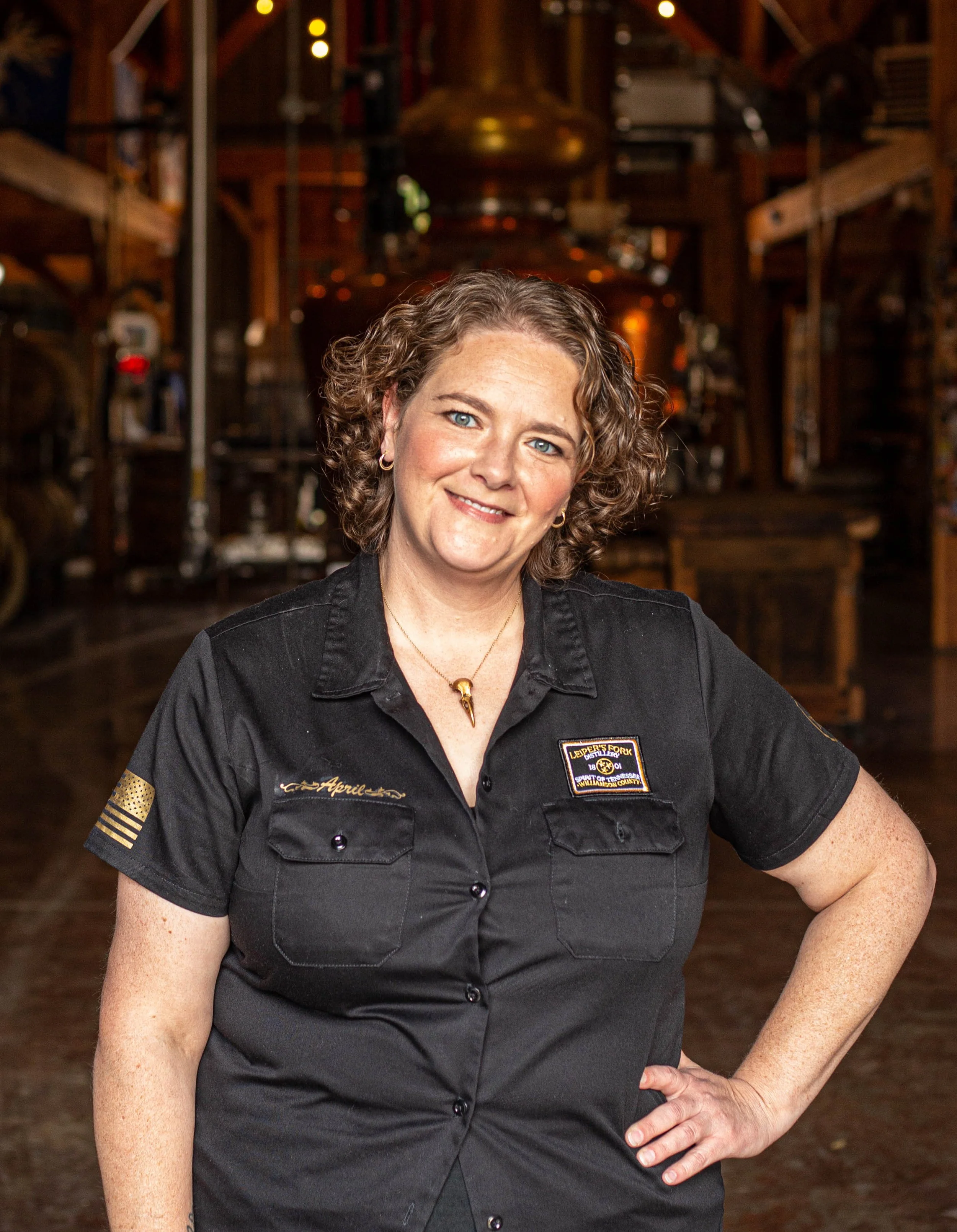 A woman with curly brown hair, wearing a uniform with patches, standing in a rustic, warmly lit interior of a brewery or similar establishment.
