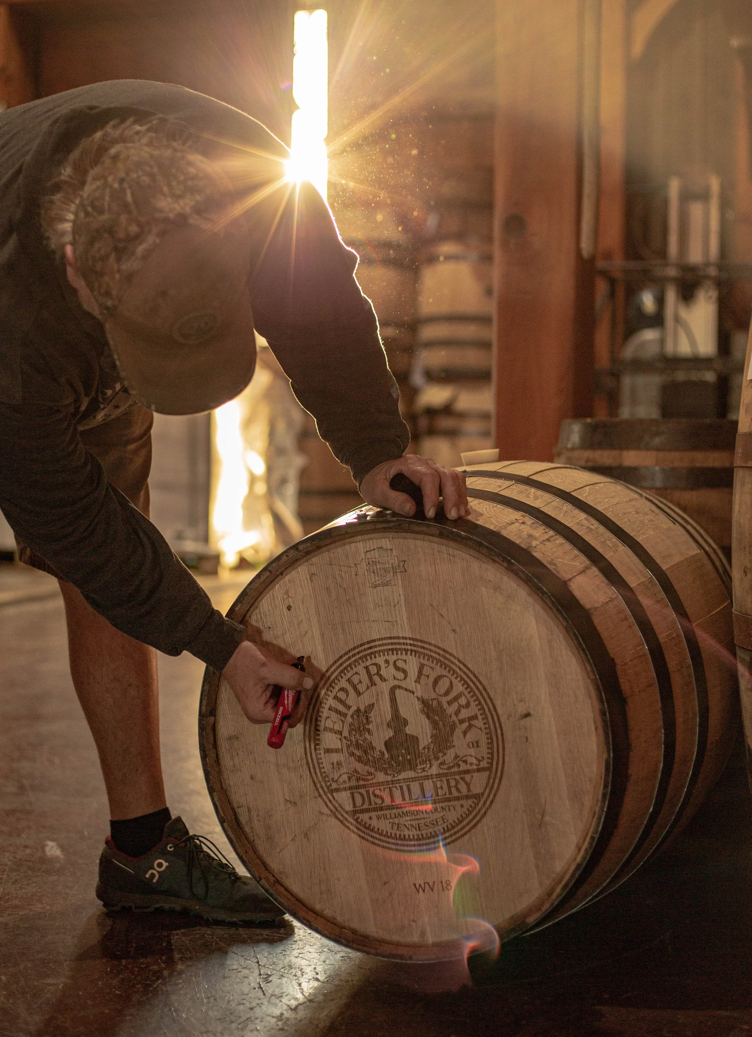 Person inspecting a whiskey barrel in a distillery with sunlight streaming through a window.