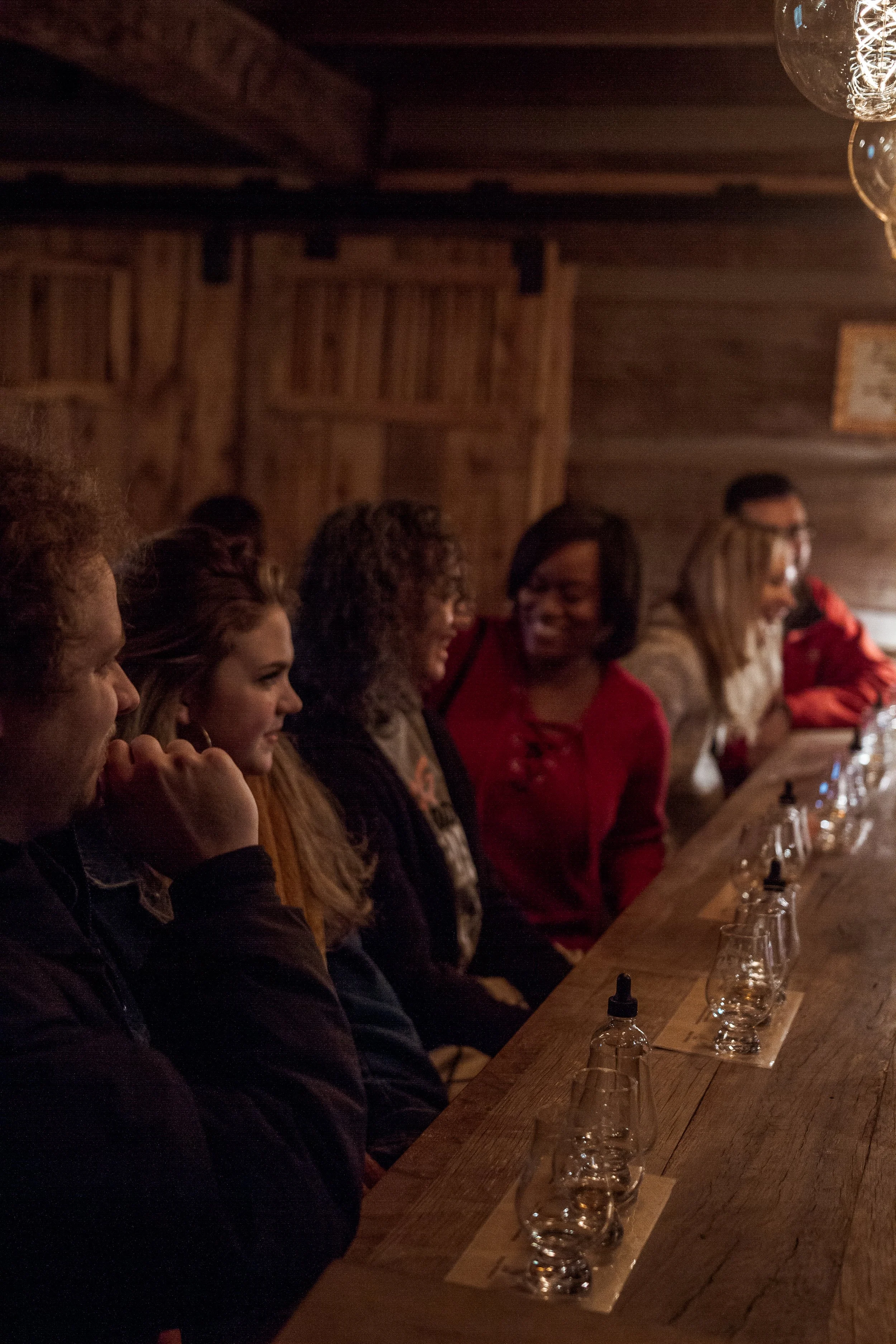A group of women sitting at a wooden bar counter in a dimly lit rustic bar or brewery, engaged in conversation.