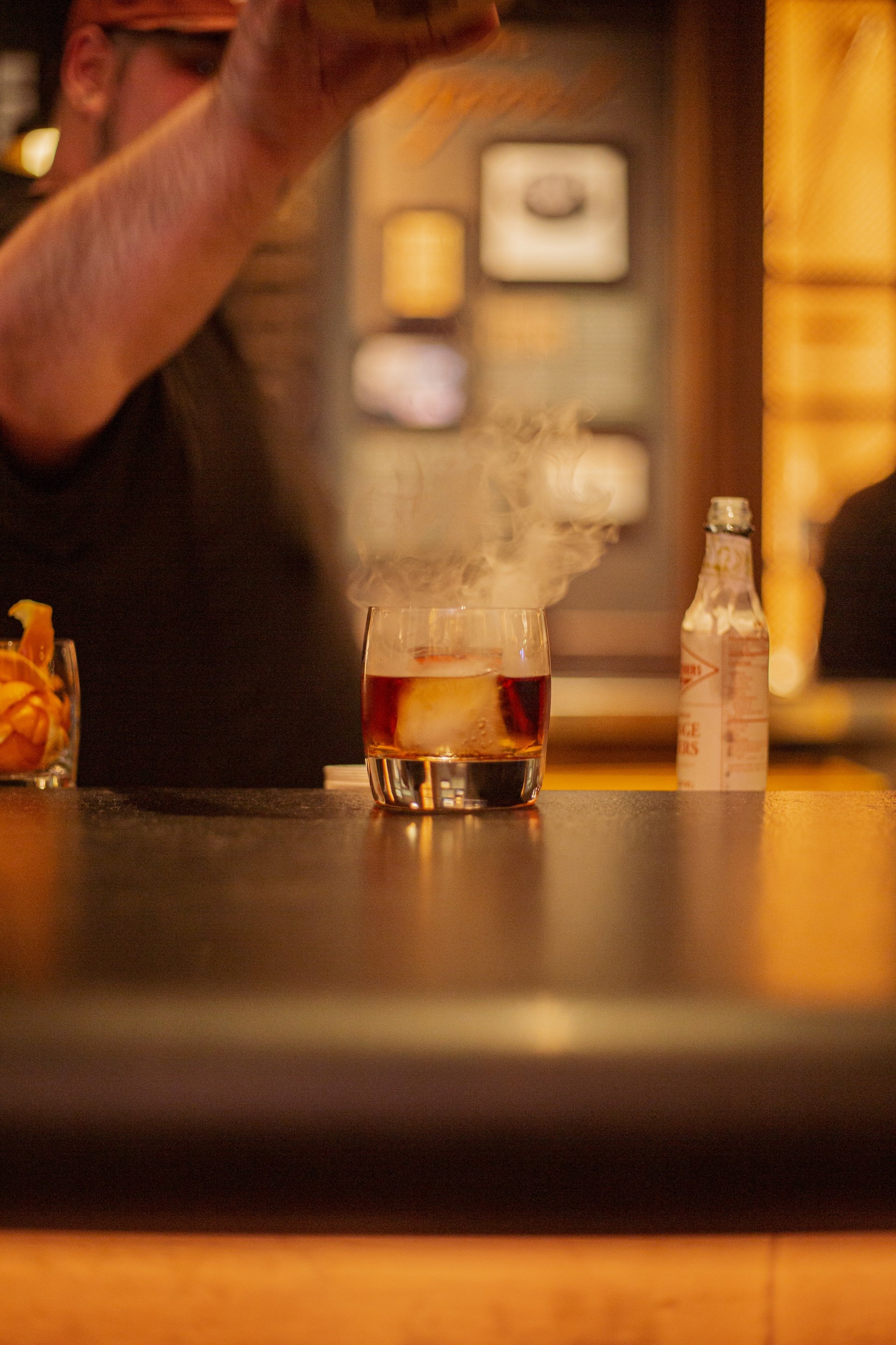A glass of whiskey with ice and steam rising from it on a bar counter, with a person behind pouring a drink and a bottle of ginger beer nearby.