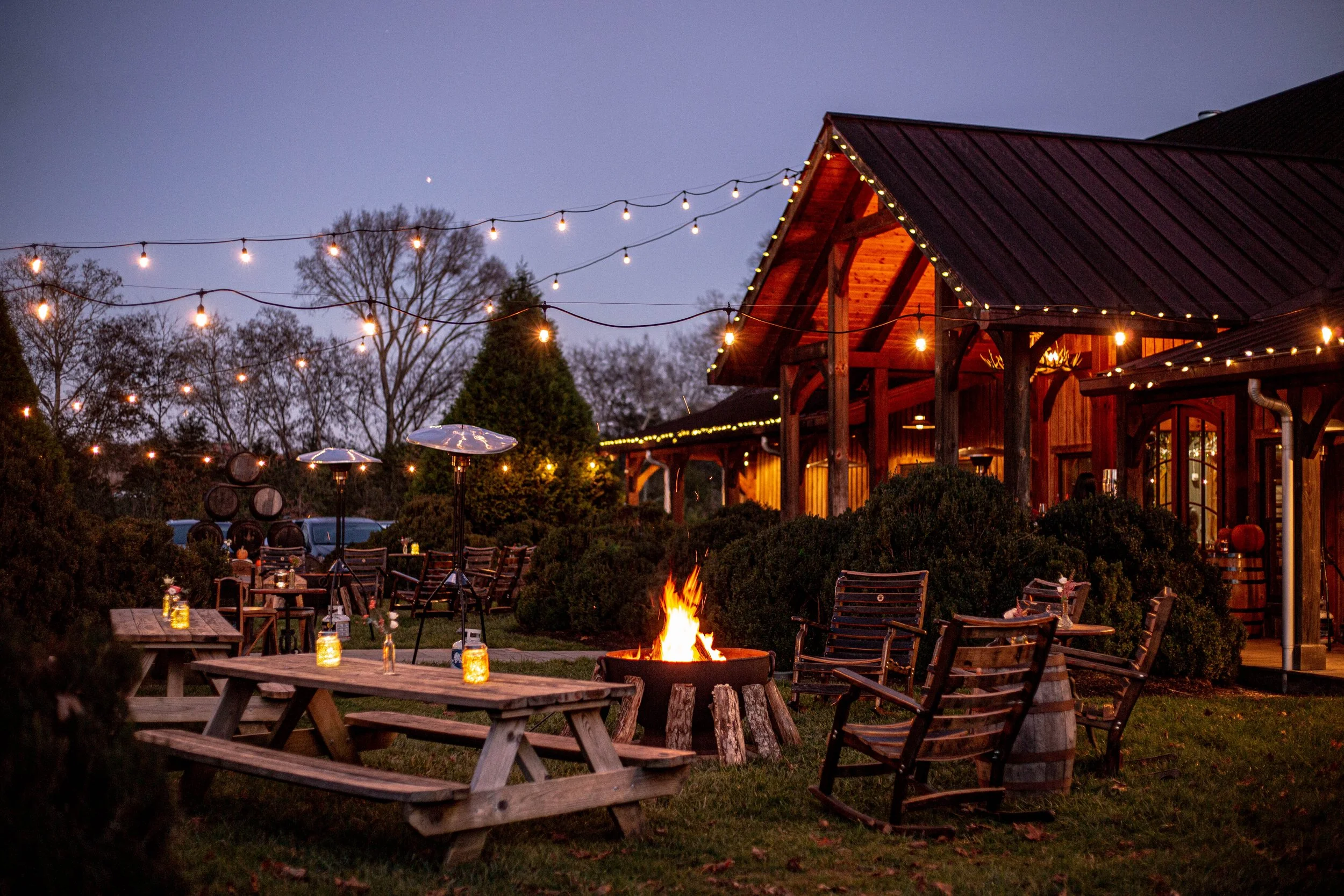 An outdoor dining area at twilight with string lights, picnic tables, chairs, and a fire pit in front of a rustic wooden building.