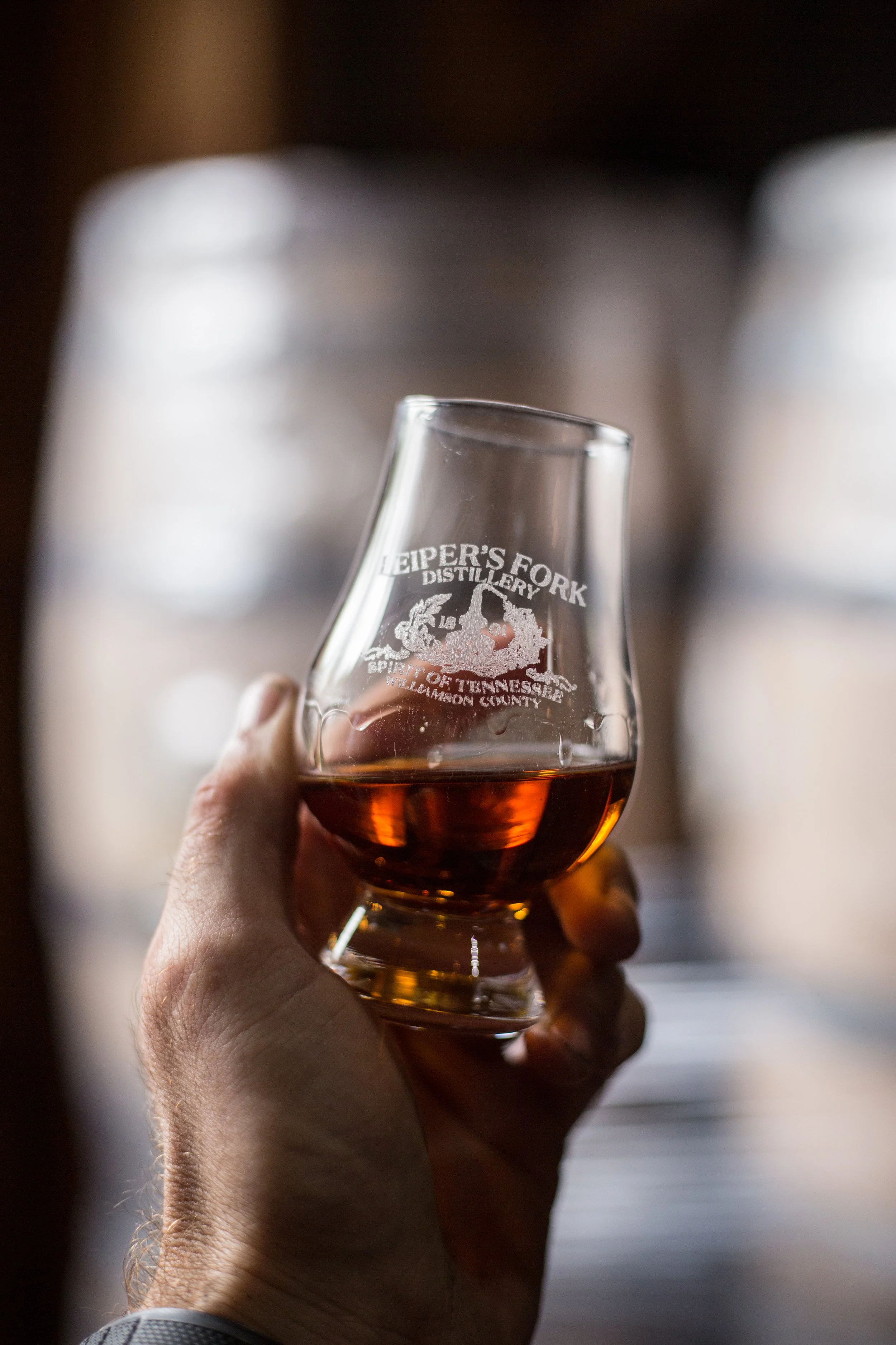 Close-up of a hand holding a glass of amber-colored liquor with the logo 'Heiper's Fork Distillery, Spirit of Tennessee, Anderson County' etched on it. The background is blurry.