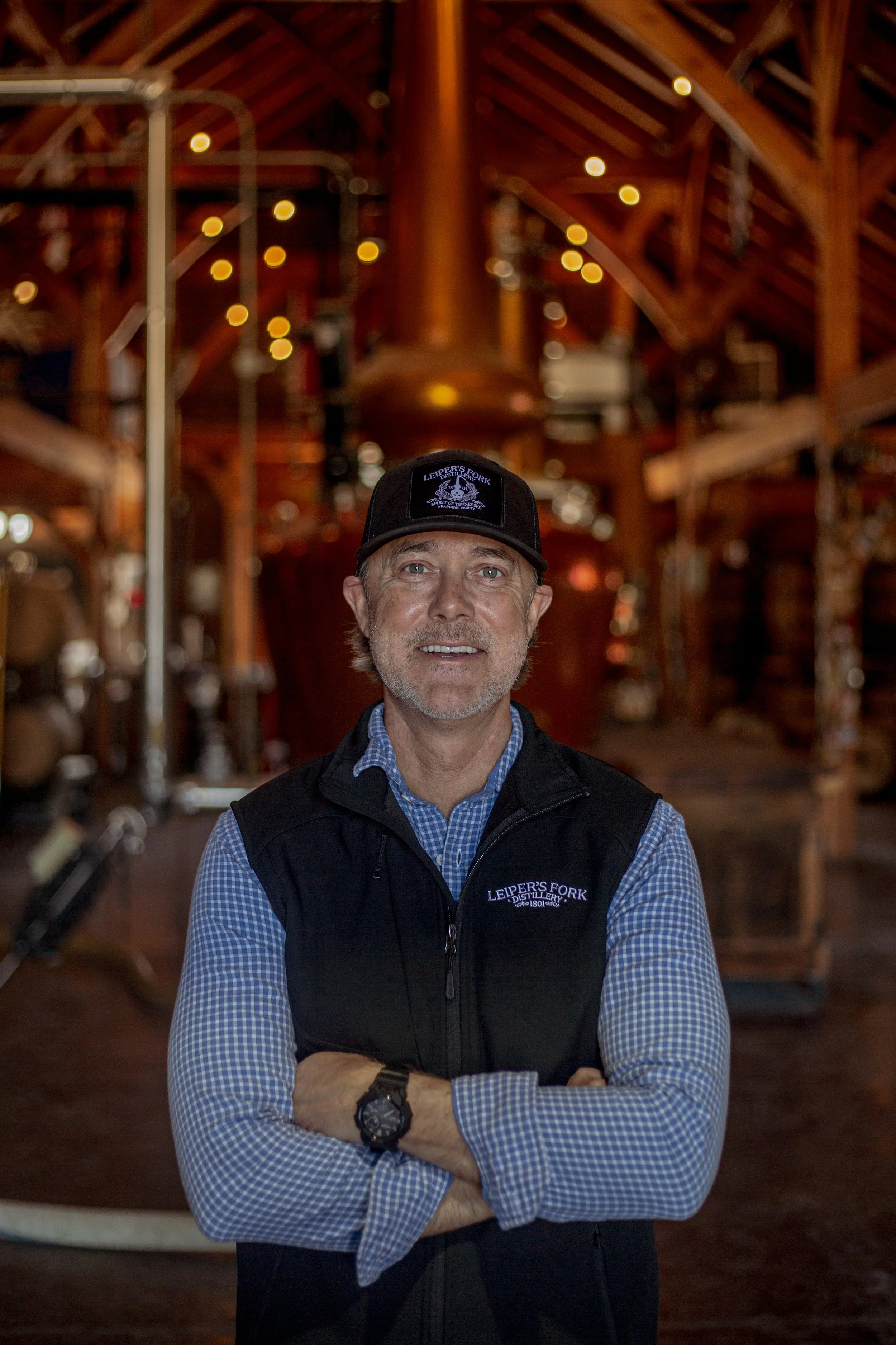 A man wearing a black vest and baseball cap with 'Leiper's Fork Distillery' logo, standing inside a rustic, wood-paneled building with string lights hanging from the ceiling, arms crossed, smiling.