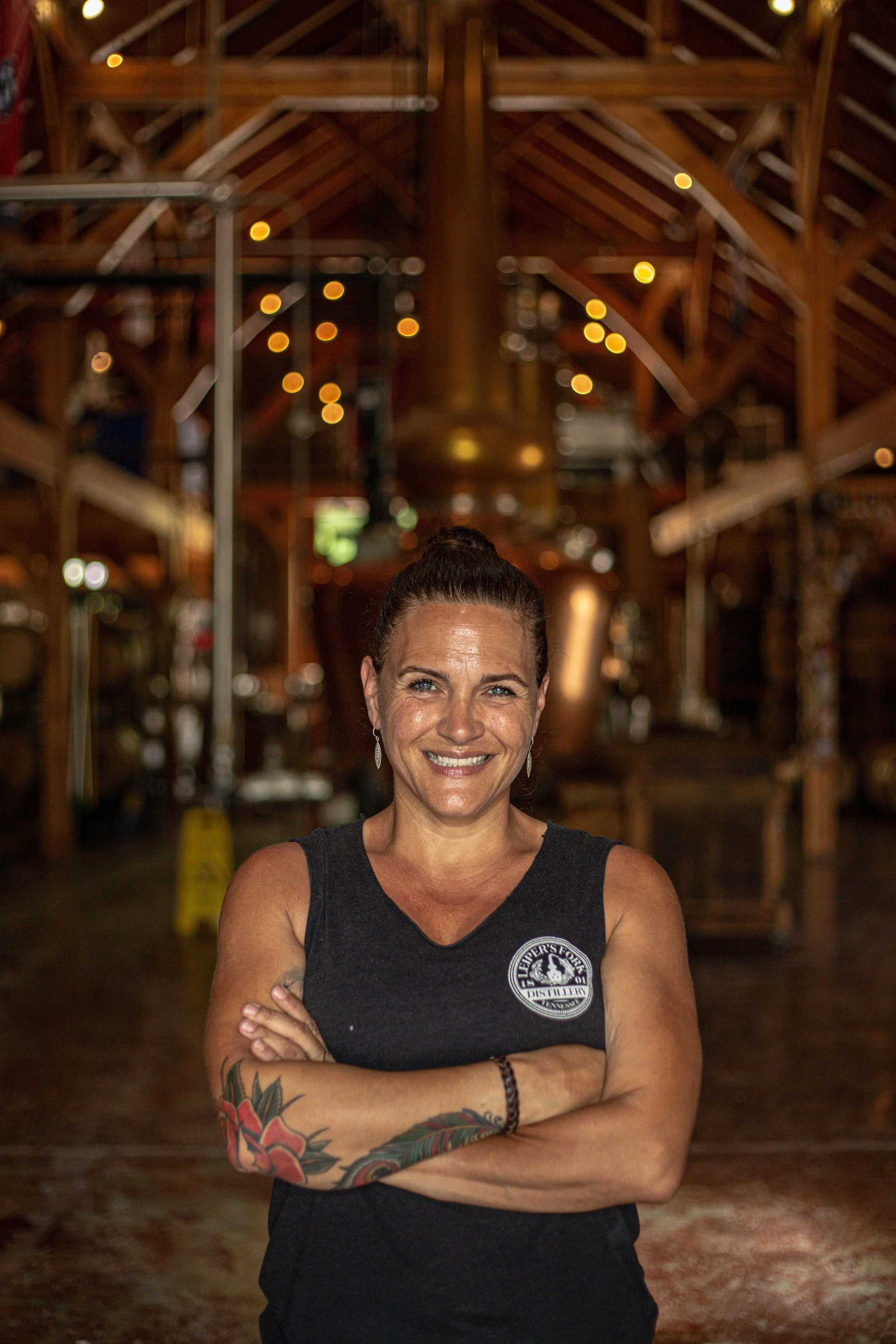 A woman with brown hair tied up in a bun, standing with arms crossed, smiling at the camera inside a wooden rustic-style venue.