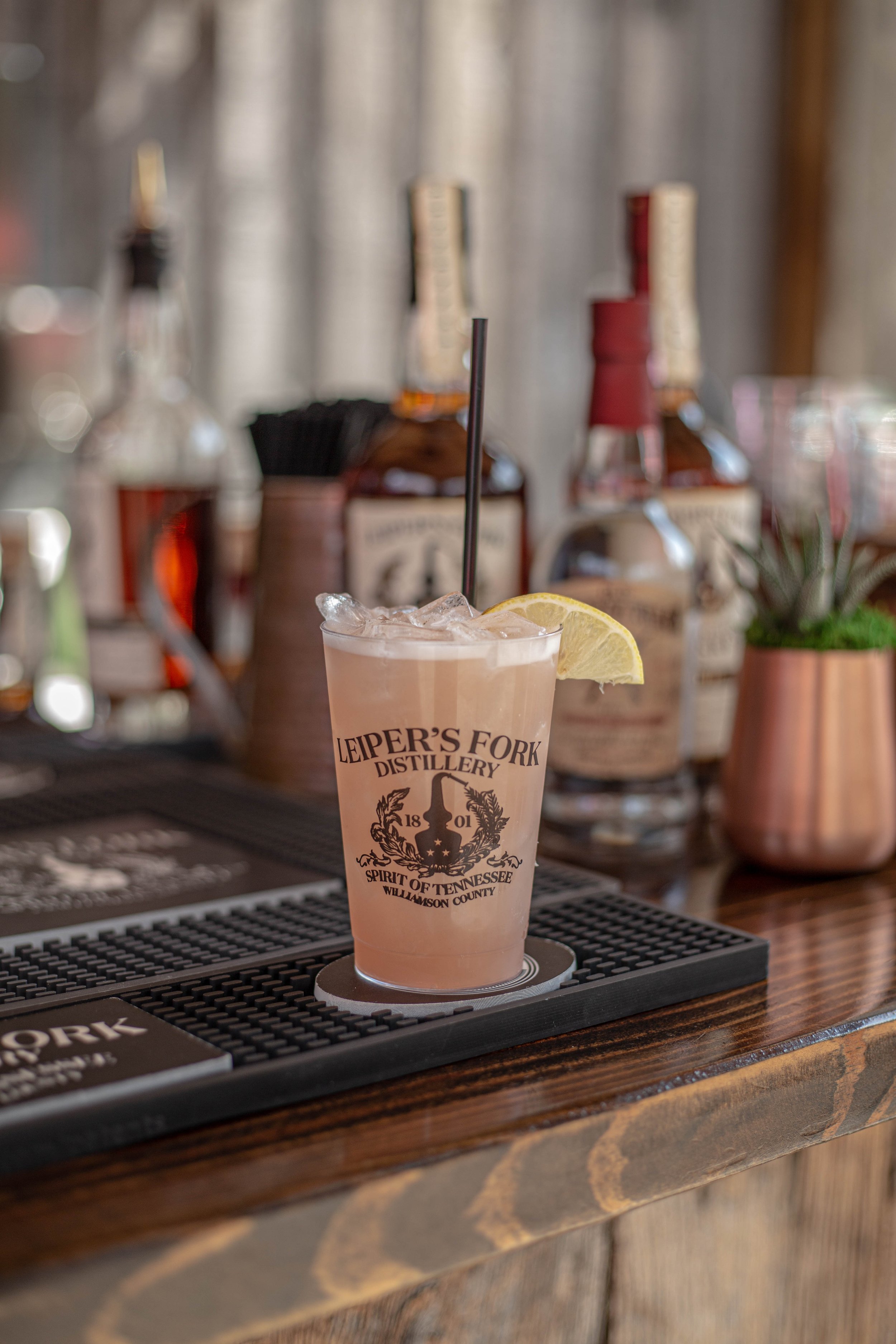A frosted cocktail with a lemon wedge garnish and a black straw on a bar counter, with bottles of alcohol and a potted plant in the background.