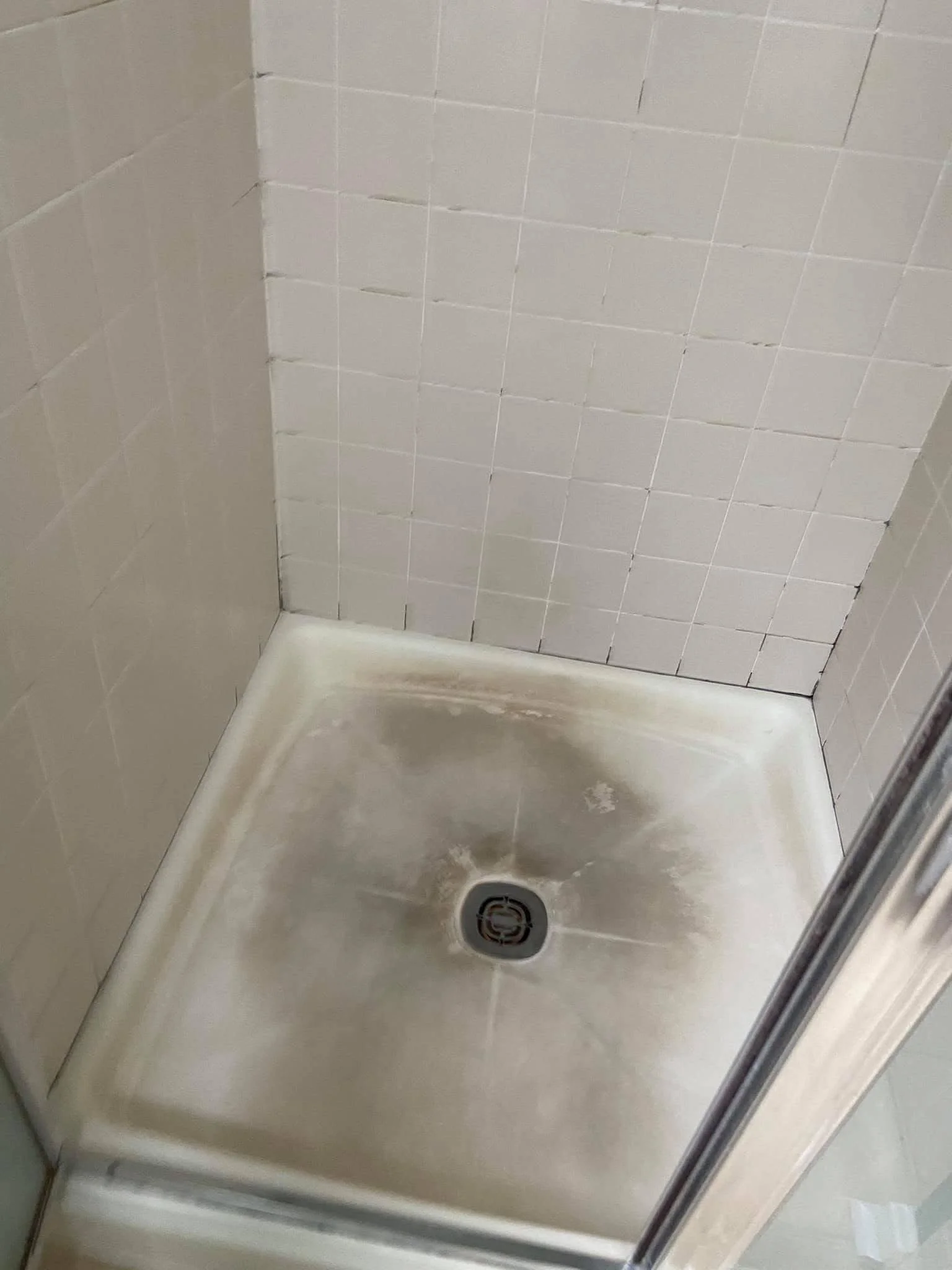A bathroom shower with white tiled walls and a white shower base, including a drain in the center.