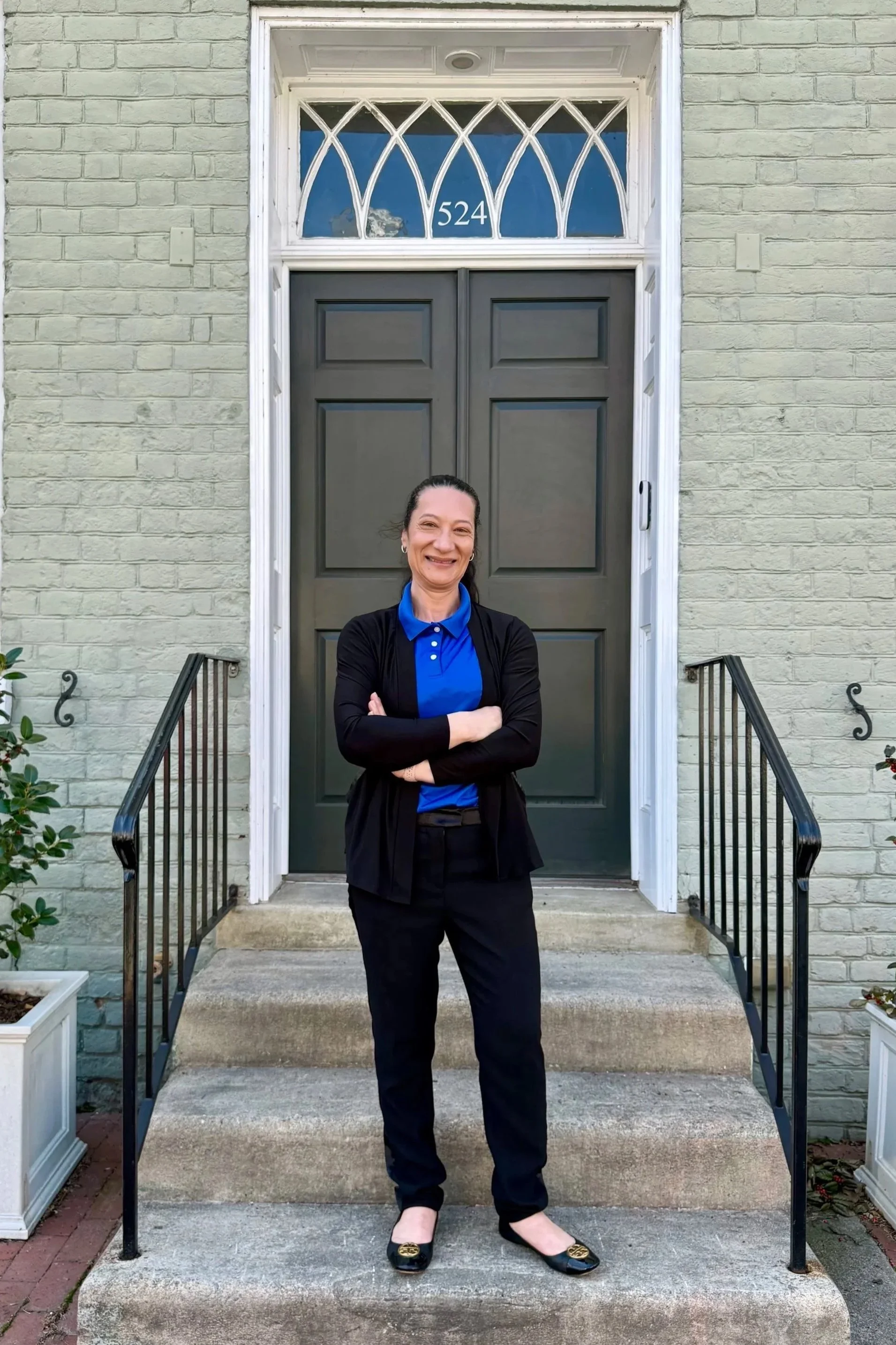 A woman standing on the steps outside a house with a gray door and a white brick exterior. She is smiling, crossing her arms, wearing a blue polo shirt, black pants, black shoes, and a black cardigan. There are small plants in white planters on either side of the steps.