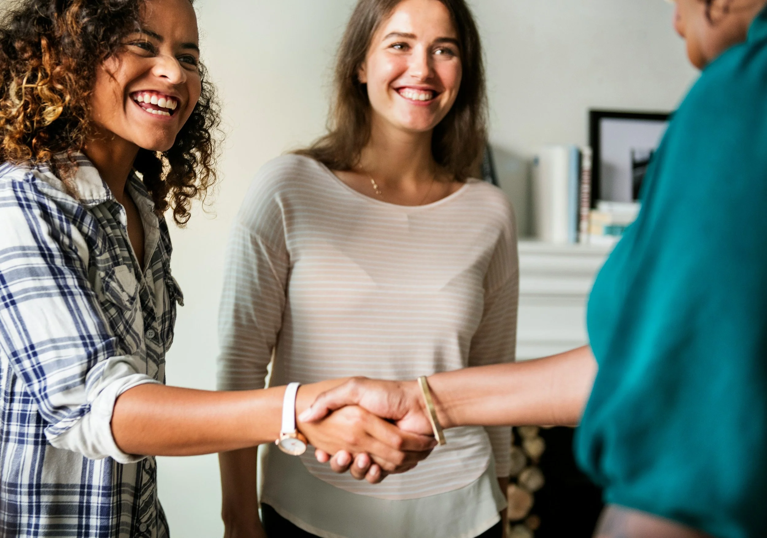 Two women smiling and shaking hands with a third person, in a professional setting.