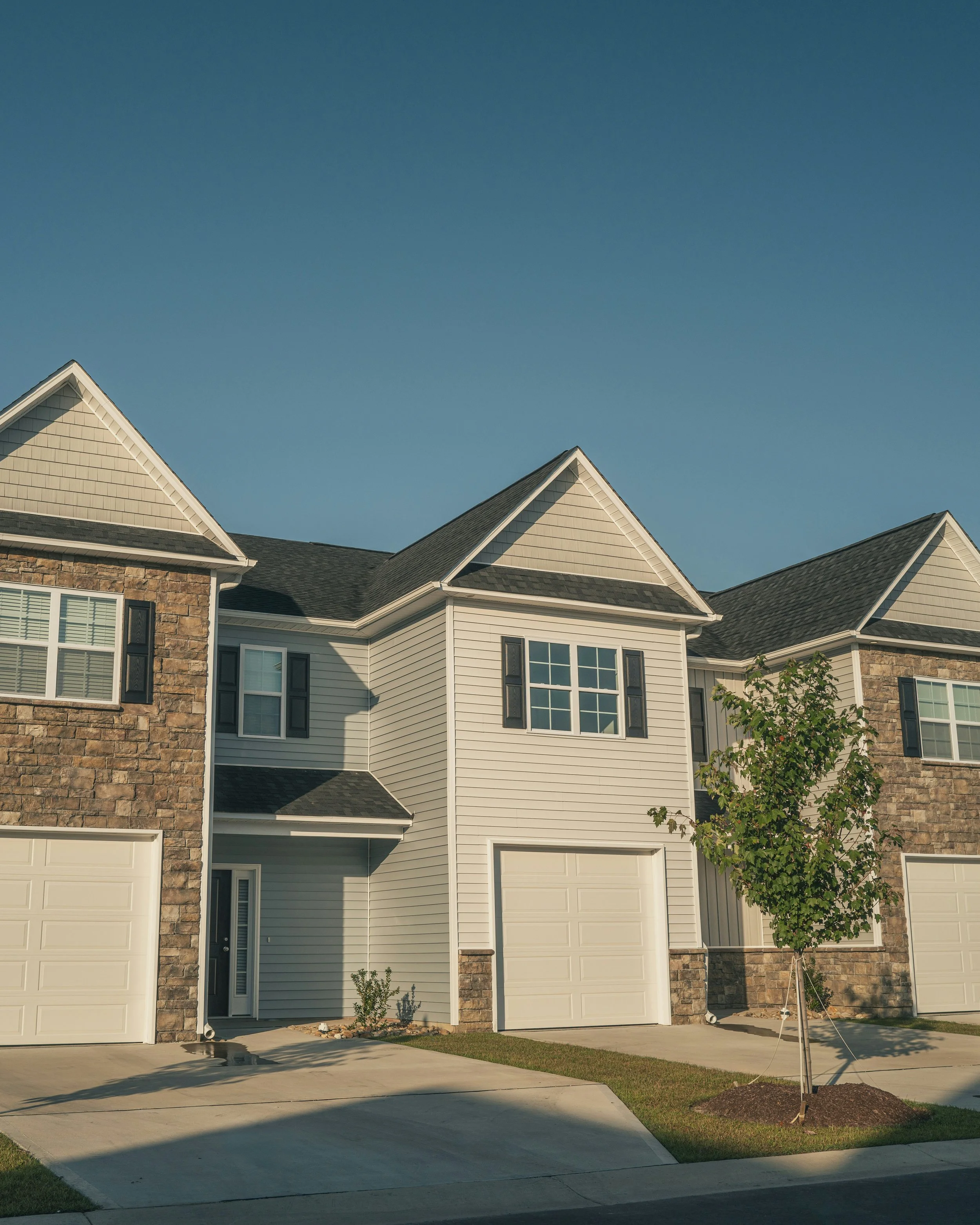 Front view of a modern residential house with beige siding and stone accents, white garage doors, a small front yard with a tree, and a clear blue sky.