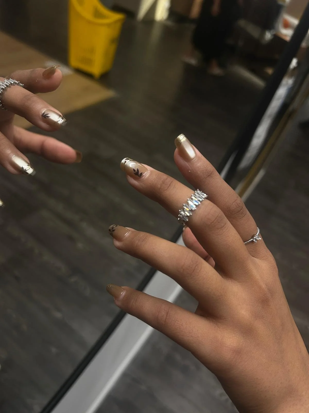 Close-up of a person's hand with gold and black decorated nails, wearing multiple rings, reflected in a mirror, with a yellow cleaning bucket and a person in black in the background.