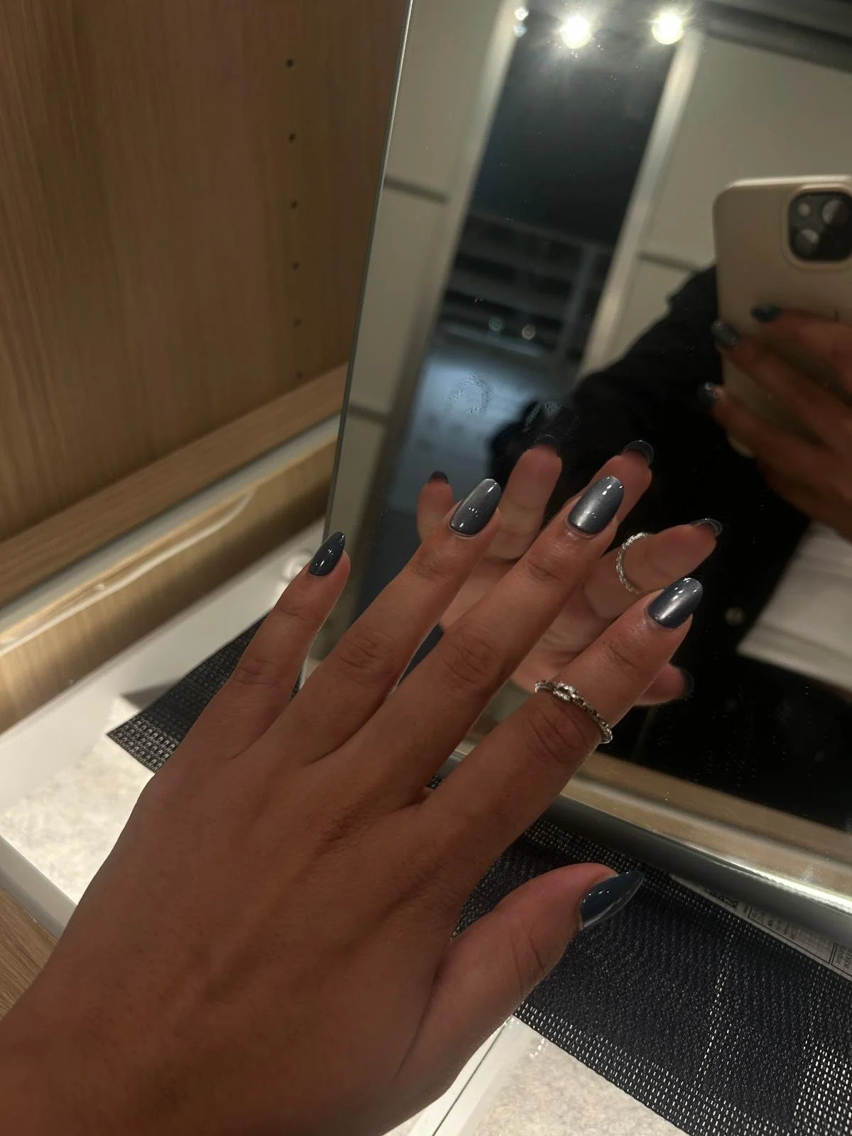 Close-up of a person's hand with manicured nails painted in a metallic cat eye blue color, wearing rings, reflected in a mirror.