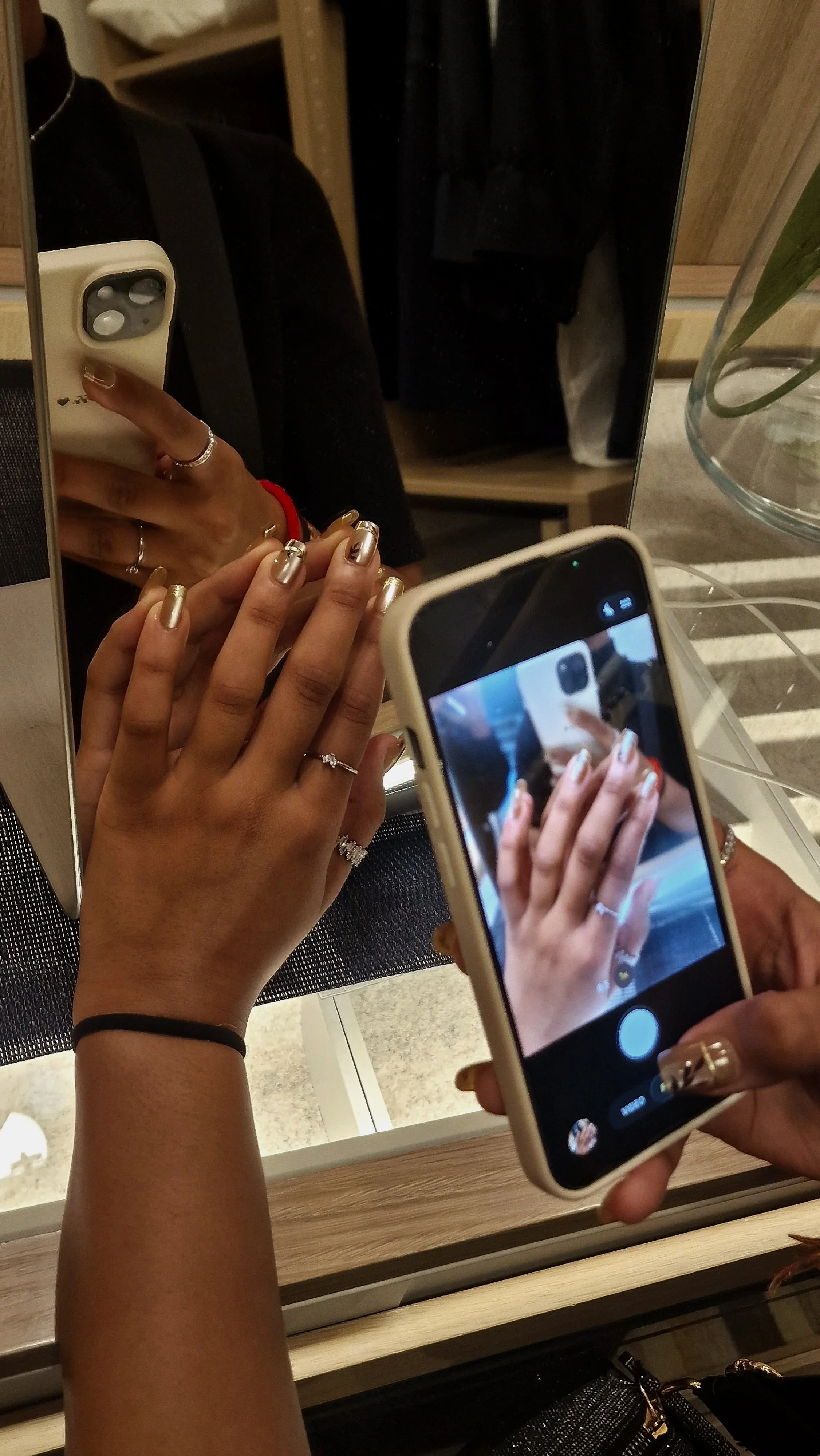 Close-up of a person's hand with metallic nail polish holding a smartphone, taking a photo of a reflection in a mirror. The reflection shows another hand with rings and silver nail polish, as well as a person's arm and part of their torso.