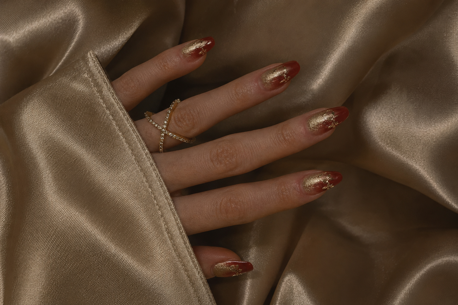A hand with gold and red metallic nail polish, wearing a delicate gold ring, resting on or holding shiny gold fabric.