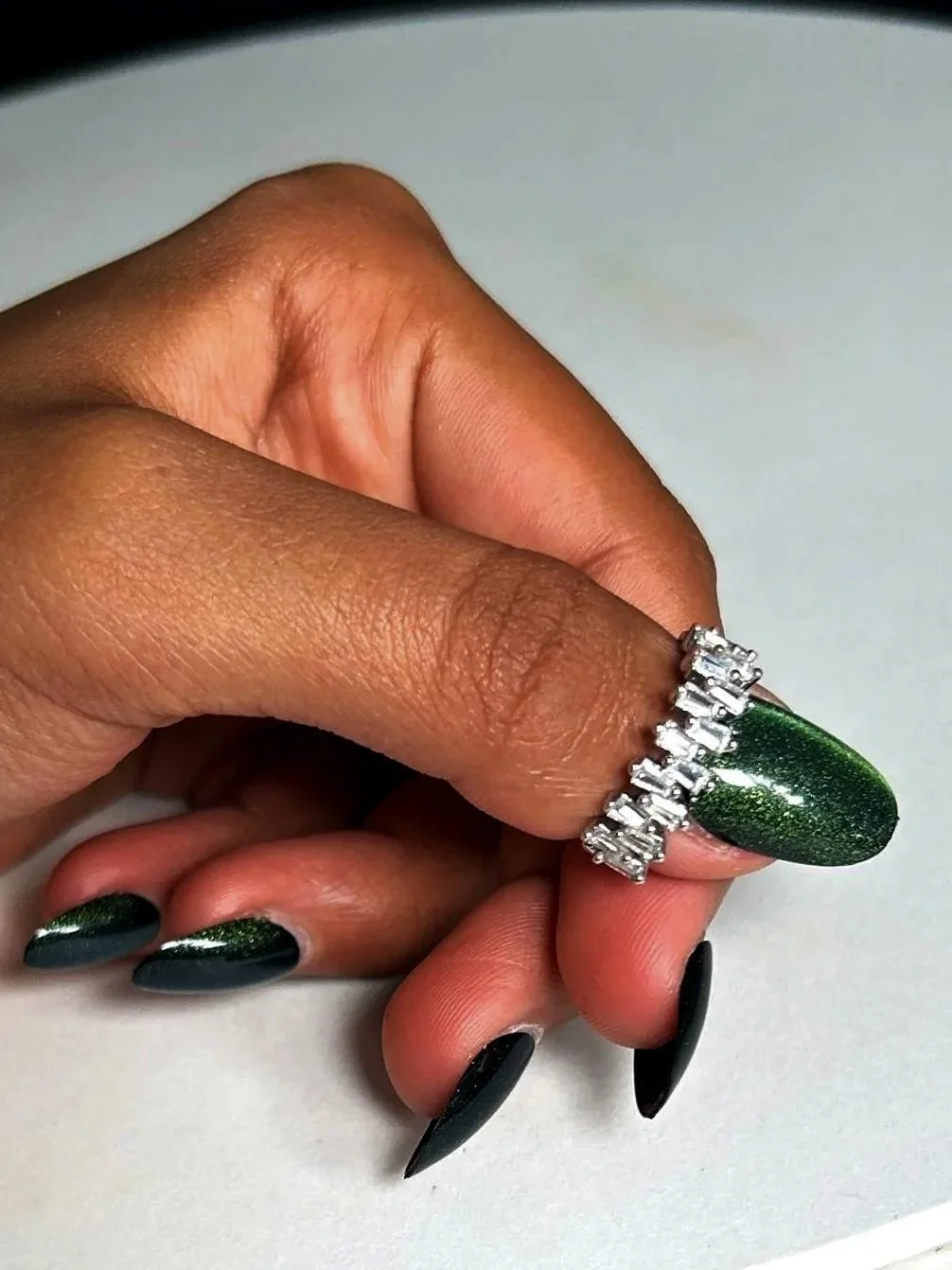 A person with brown skin is posing with their fingers, which have dark green metallic cat eye nail polish, wearing a silver ring on thier thumb