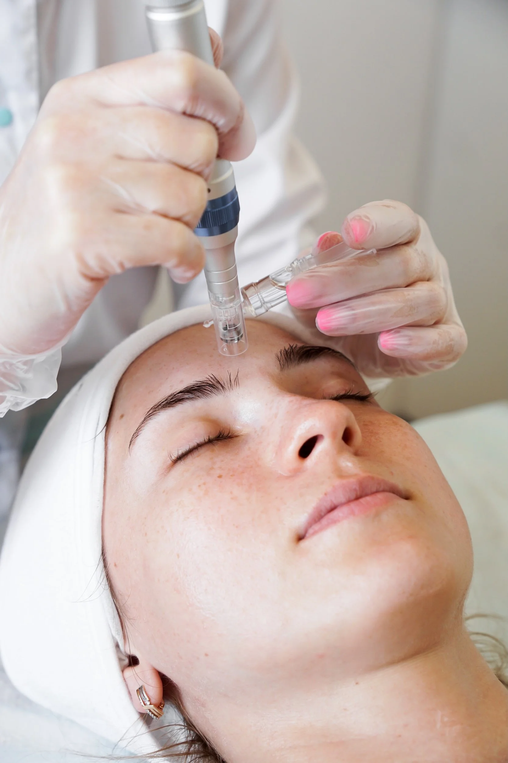 A person lying down with their eyes closed, receiving a facial treatment with a skincare device applied to their forehead, in a clinical setting.