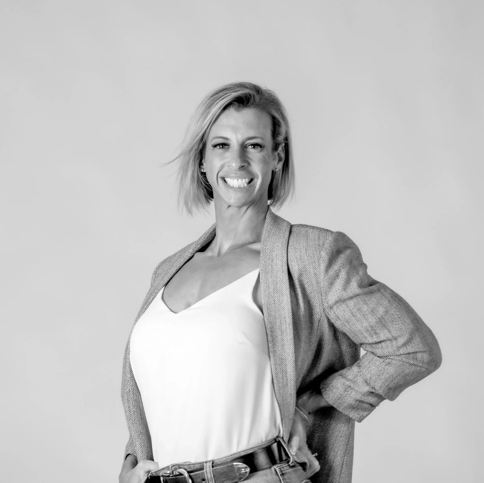 A black and white portrait of Jessica with a bright smile and shoulder-length hair. She is wearing a textured blazer draped over her shoulders, a light-colored top, and a belt, posing confidently against a neutral background.