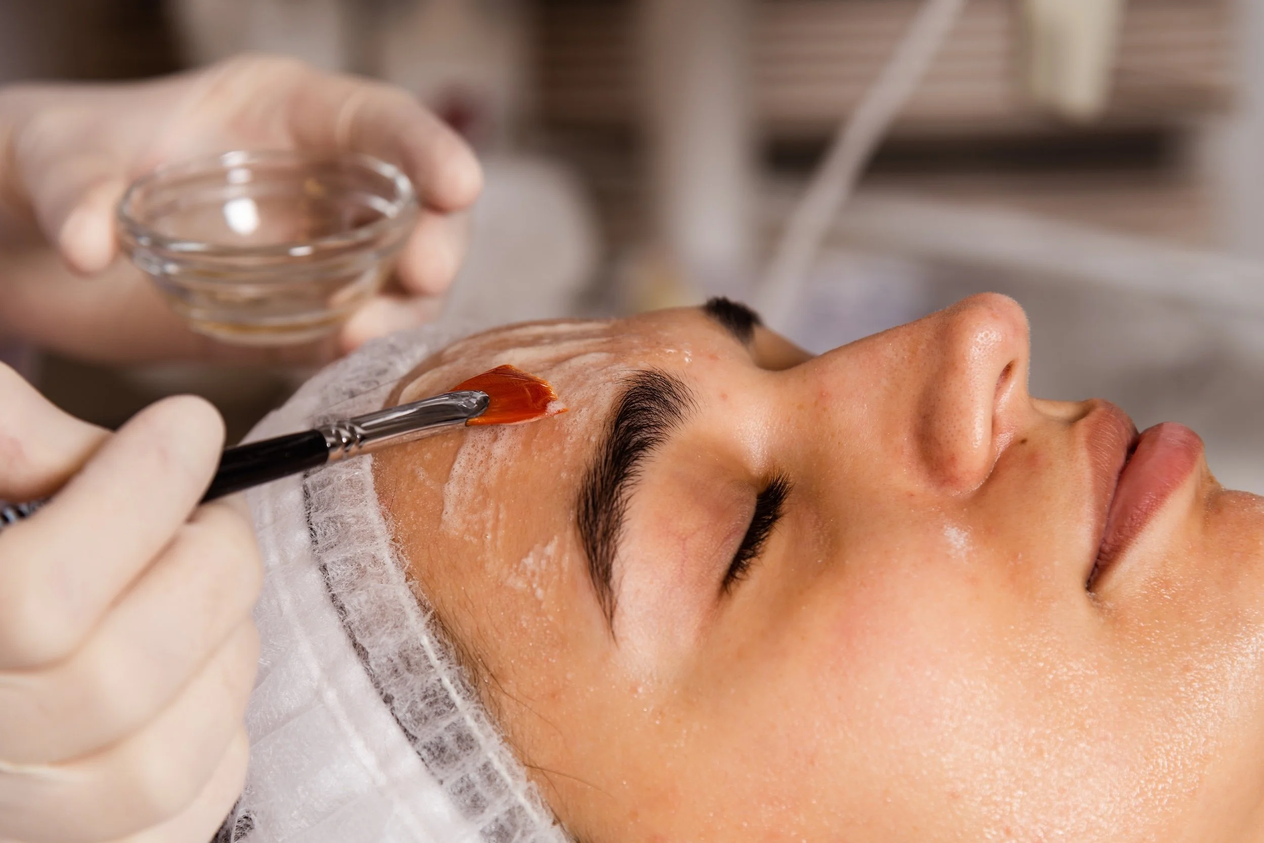 A person receiving a facial treatment in a spa or skincare clinic. A beautician is applying a skincare product with a brush on the client's forehead while wearing gloves. The client has closed eyes and appears relaxed.