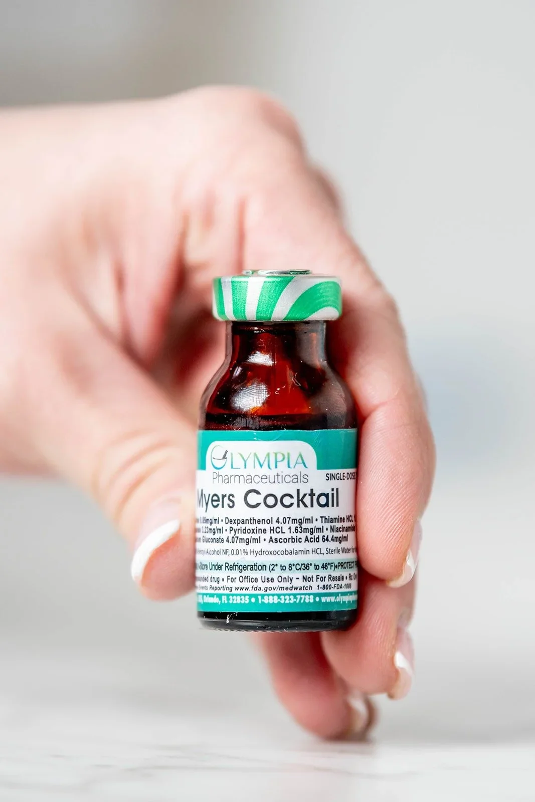 A close-up shot of a hand holding a single-dose glass vial labeled "Myers Cocktail." The label lists ingredients such as Magnesium Chloride, Dexpanthenol, Thiamine, and Ascorbic Acid. The vial has a distinctive teal and white striped cap.