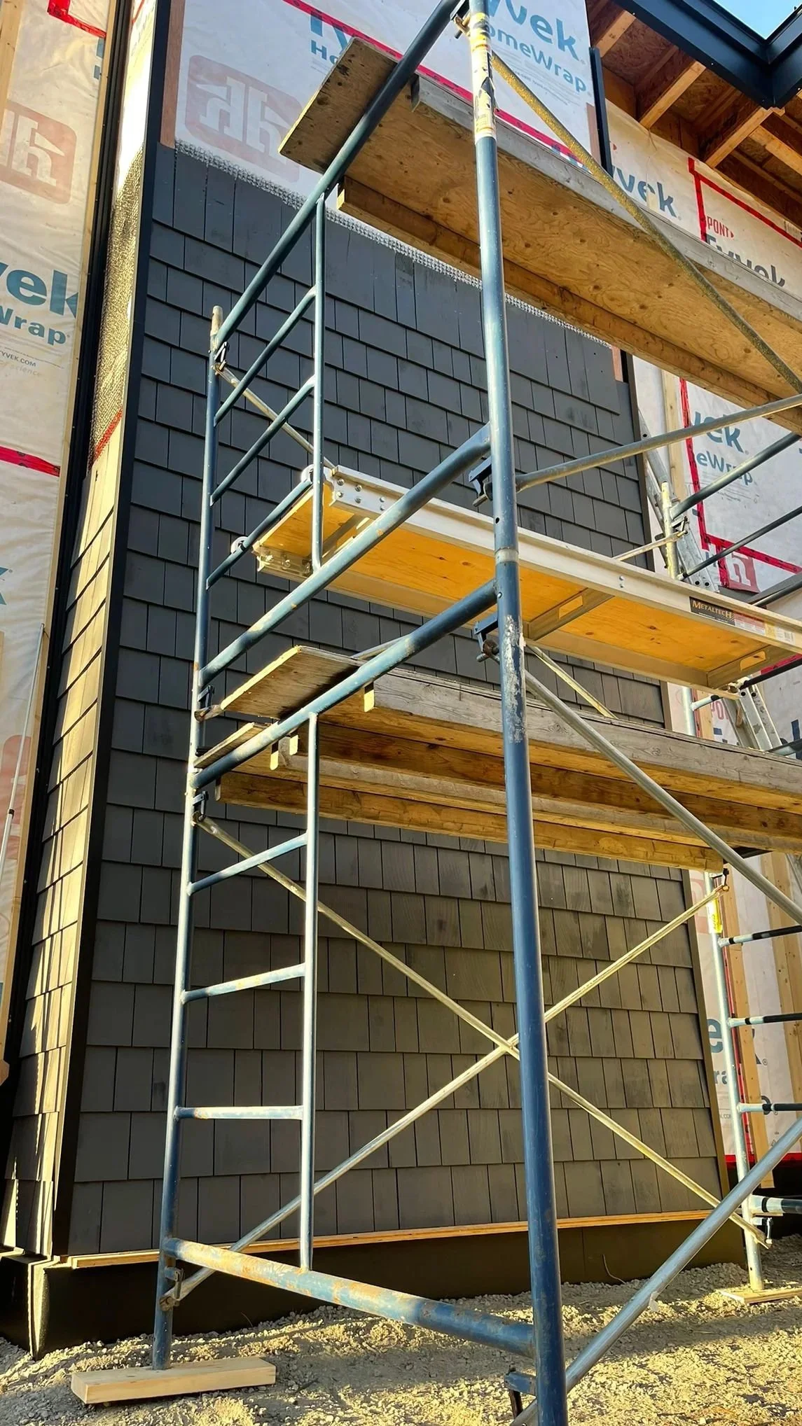 Construction scaffolding next to a building with dark gray siding and partially installed black shingles.