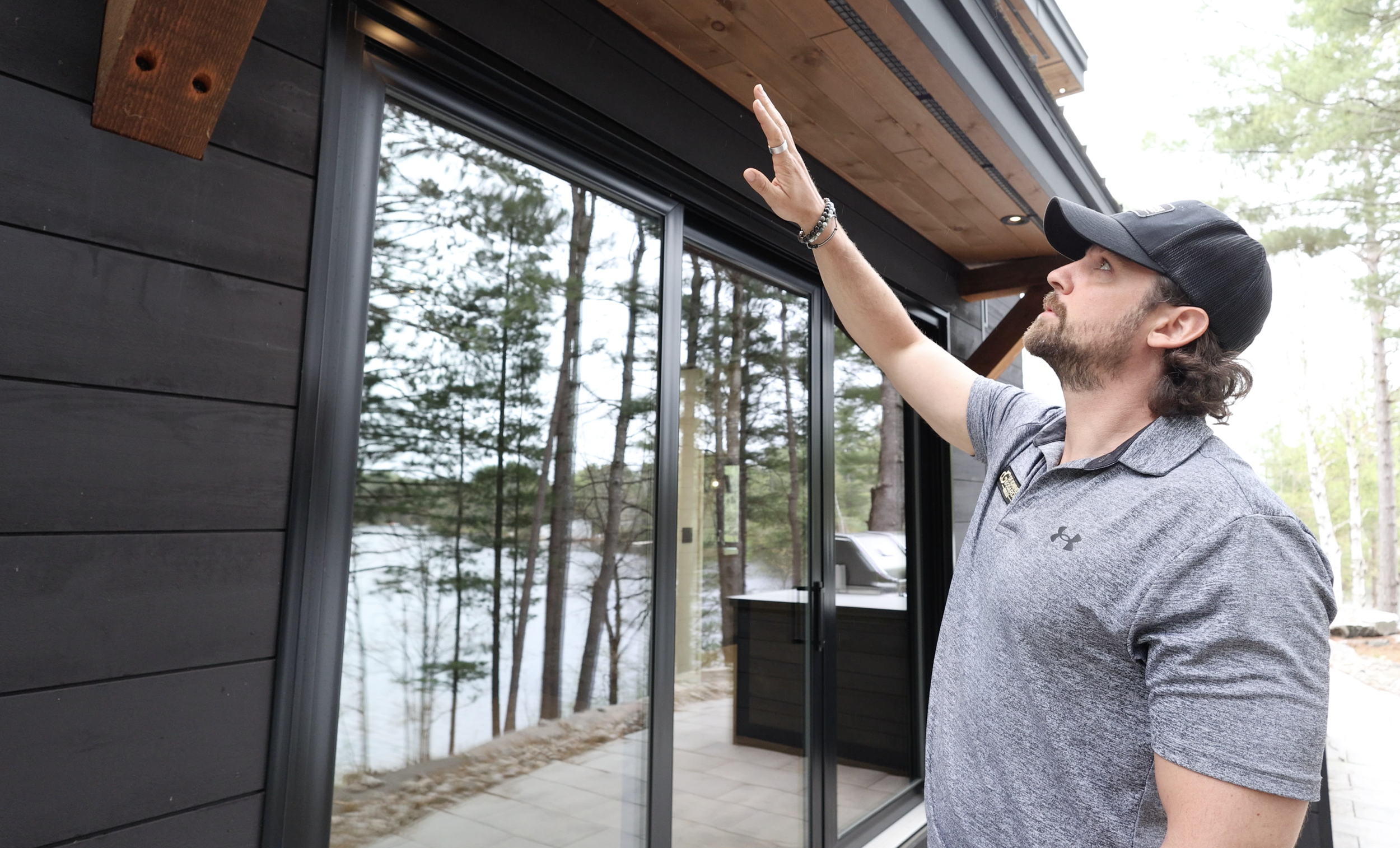 A man in a gray shirt and black cap pointing at the ceiling in front of a large glass sliding door, with trees and a lake visible outside.