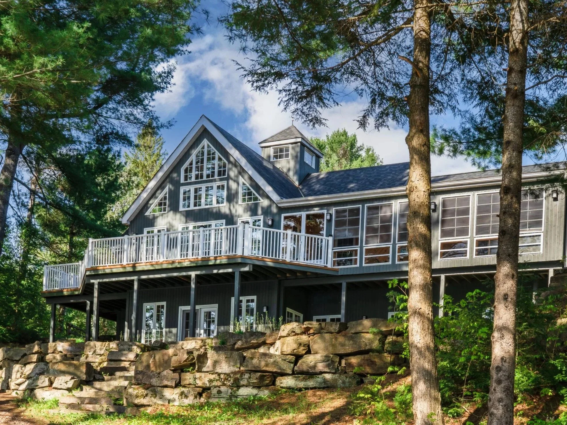 A multi-story house with a gray exterior, large windows, and a spacious white railing deck, surrounded by tall trees and natural greenery.