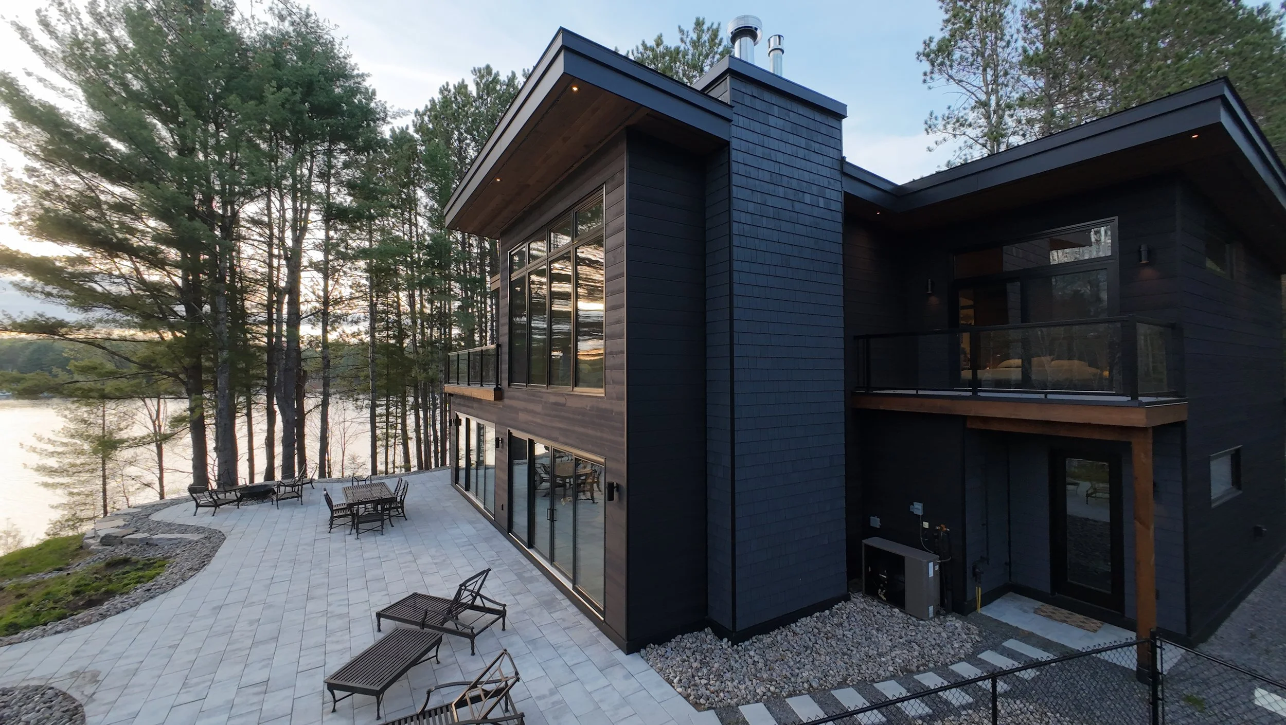 Modern black house with large windows and a balcony, surrounded by trees and overlooking a lake, with an outdoor patio area with chairs and tables.