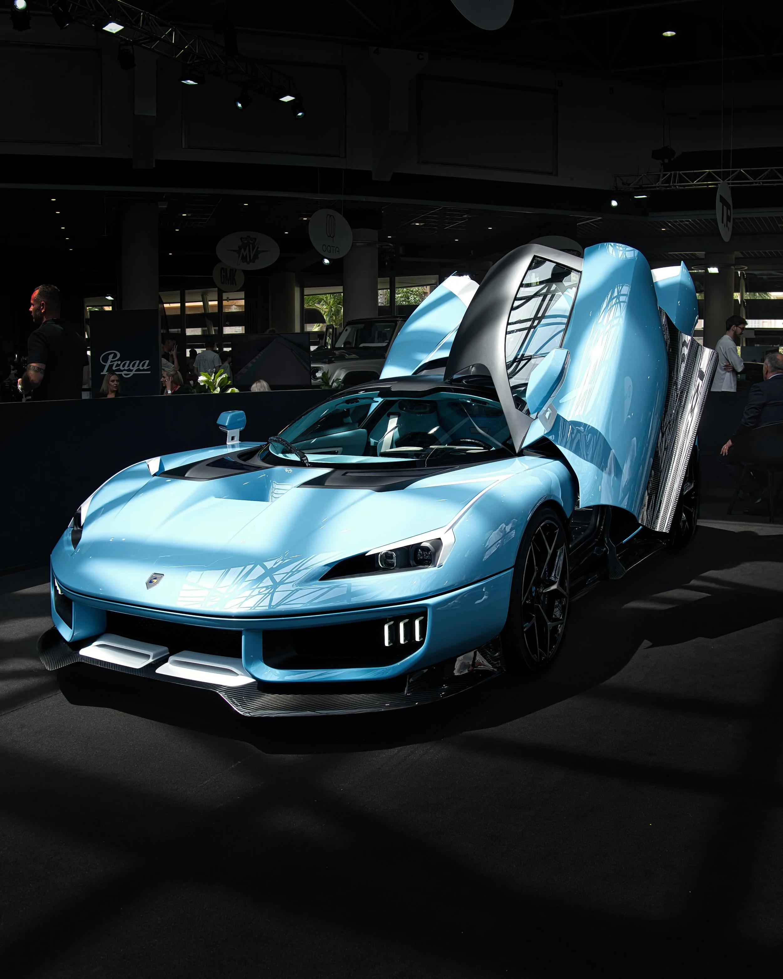 Une voiture de sport bleue avec des portes en élytre ouverte, exposée dans un salon automobile avec plusieurs personnes en arrière-plan.