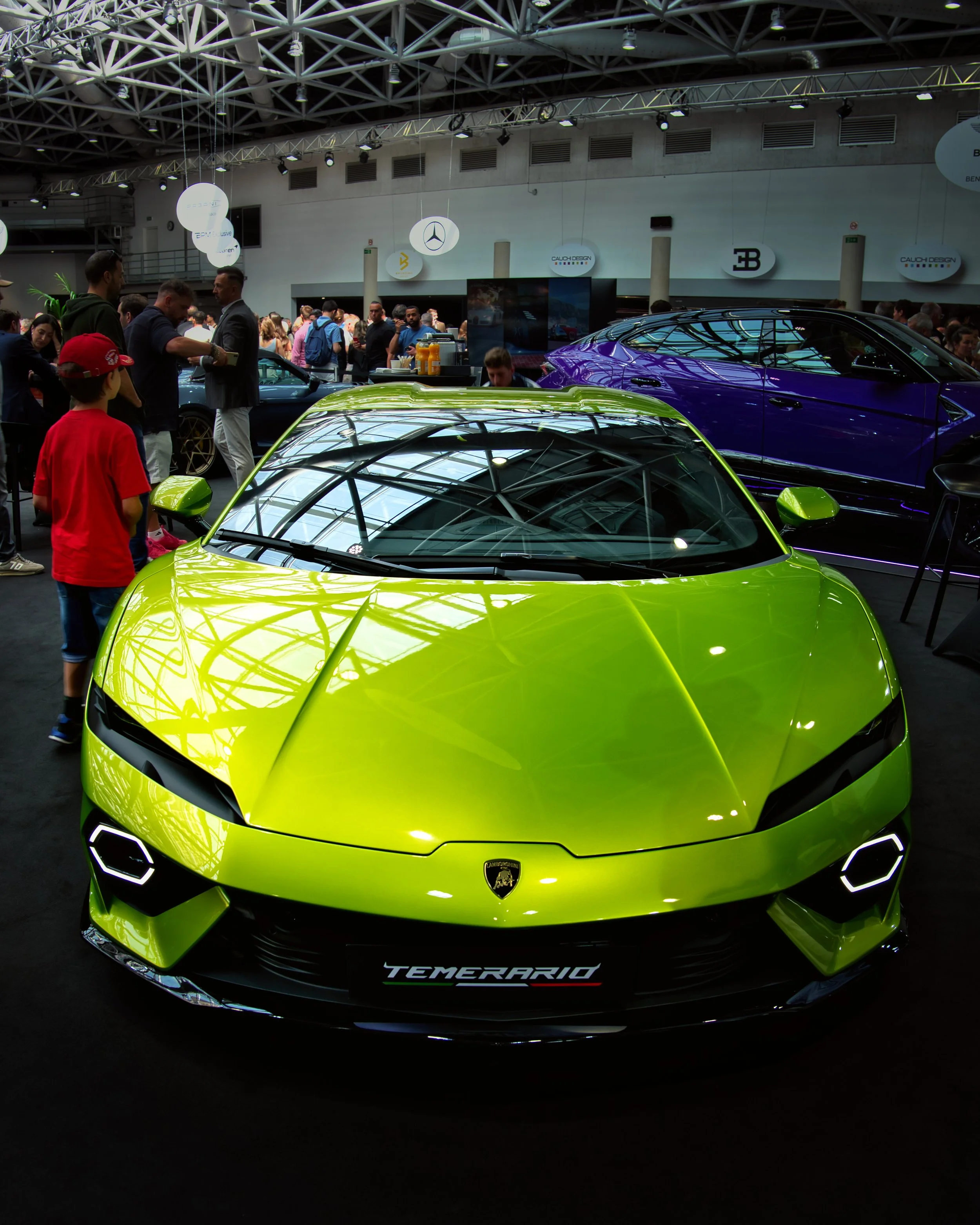 Une voiture de luxe vert lime Lamborghini, exposée lors d'un salon automobile, avec un logo en forme de bouclier sur la calandre, nommée 'Temerario'. En arrière-plan, diverses personnes admirent les véhicules et des panneaux publicitaires sont suspendus au plafond de l'exposition.