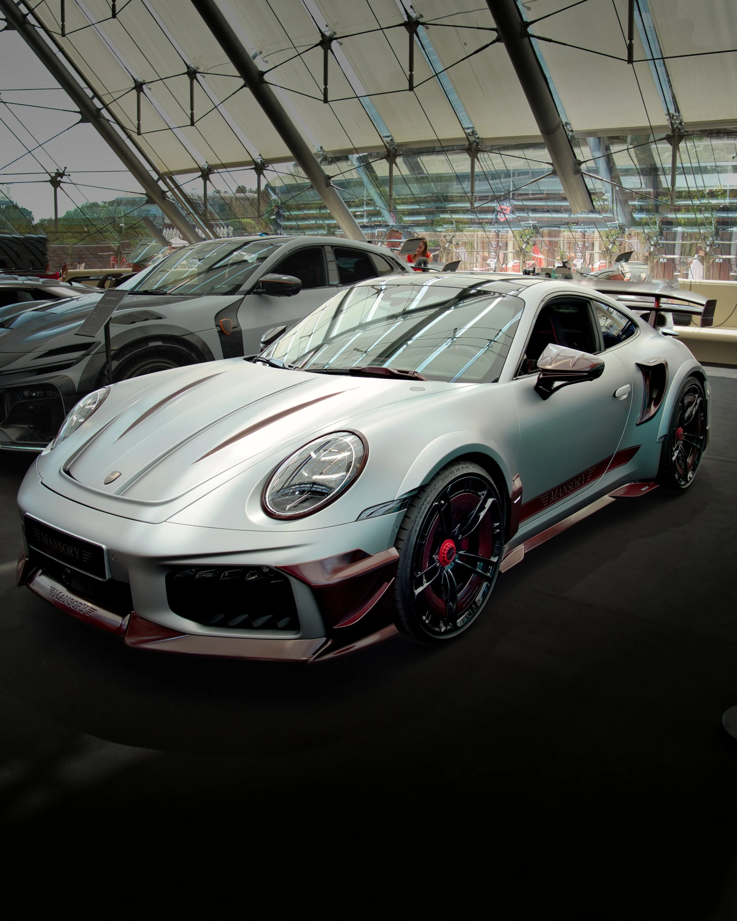 Une salle d'exposition de voitures de luxe avec plusieurs voitures de sport modernes, y compris une Porsche blanche en premier plan, dans un bâtiment avec un toit en verre.