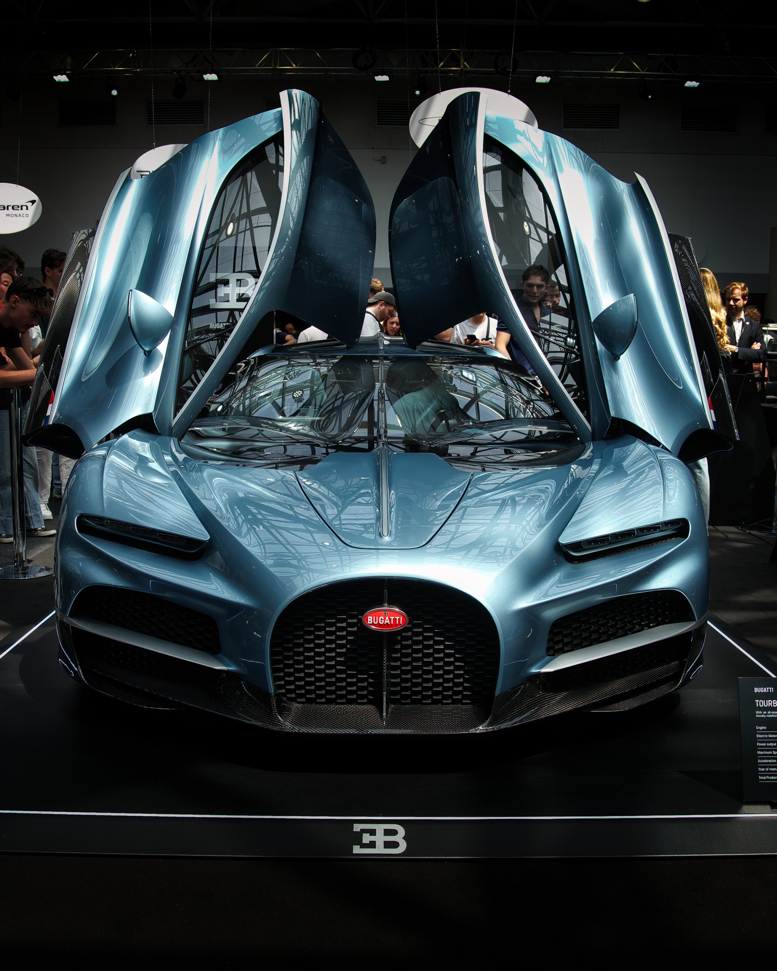 Une supercar bleue exposée lors d'un salon automobile, avec des portes en élytre en position ouverte, entourée de spectateurs.
