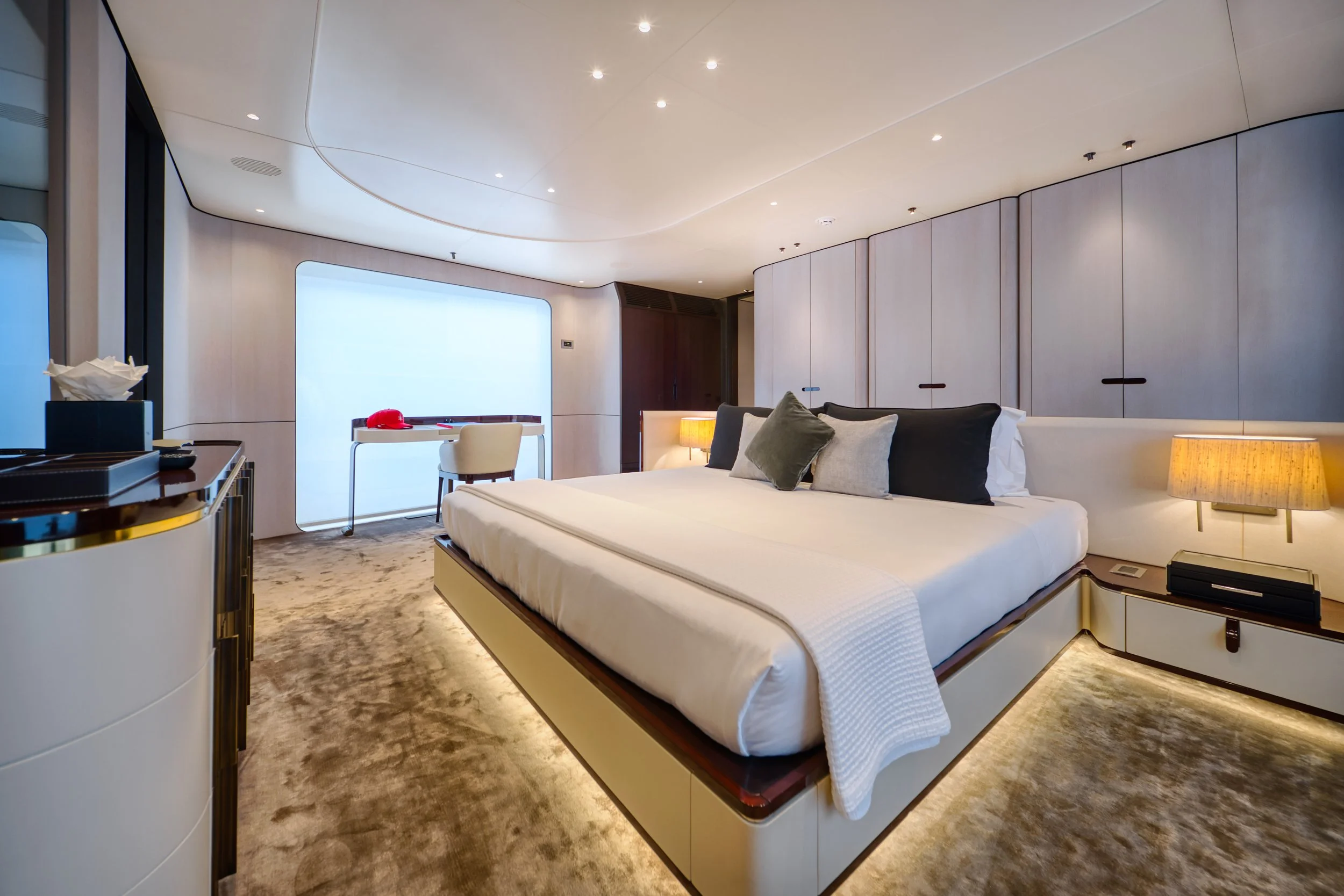 Chambre moderne avec un lit à plate-forme, coussins, chevets avec lampes, murs en placage clair, tapis beige, grande fenêtre avec voilage, meubles en bois foncé, design épuré et éclairage discret.