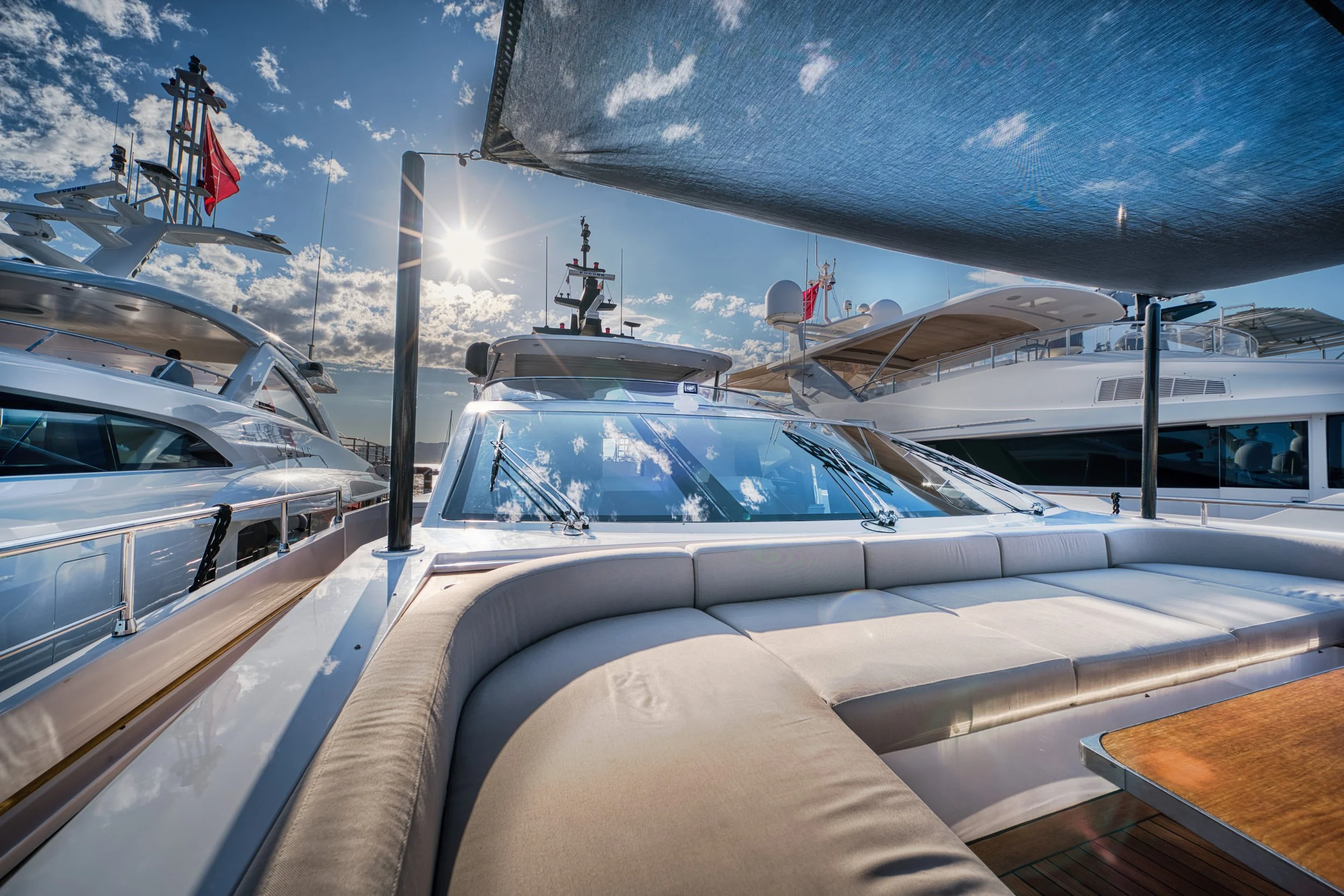 Vue d'un yacht avec un canapé confortable en plein air, plusieurs autres yachts en arrière-plan, un ciel ensoleillé avec quelques nuages et le soleil brillant.