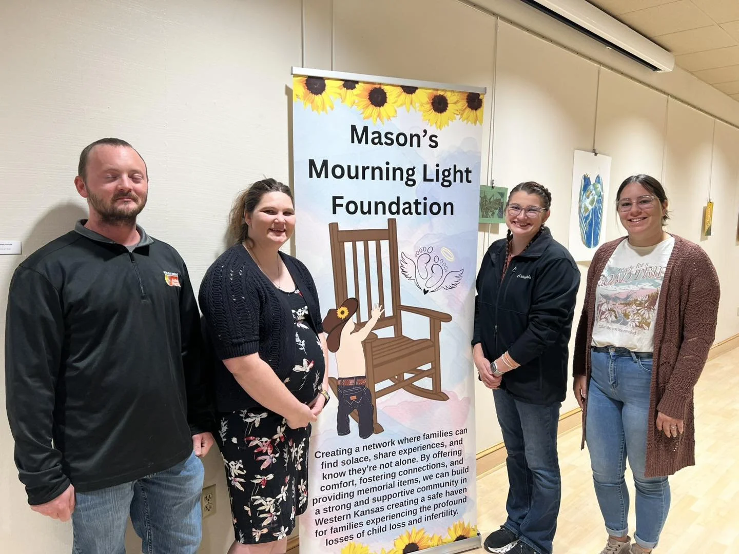Four people standing next to a banner for Mason's Mourning Light Foundation, which features a angel with wings, sunflowers, and a grieving girl.