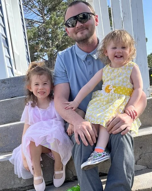 A man sits on outdoor steps with two young girls, one on each knee, smiling with trees and a railing in the background.