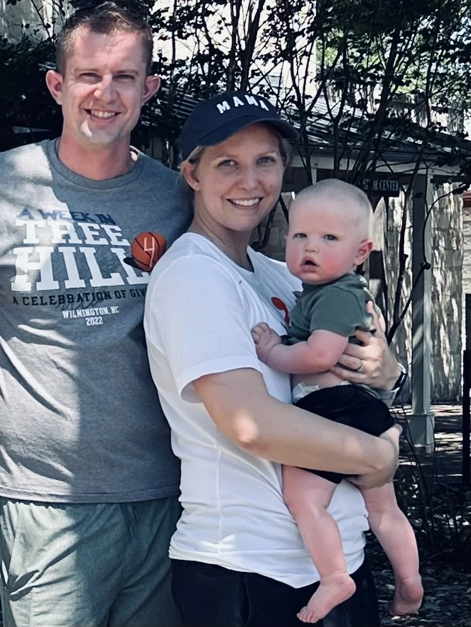 A woman holding a young child in her arms, with a man standing beside them smiling outdoors during daytime. The woman wears a cap and a white T-shirt, the child wears a green T-shirt, and the man wears a gray T-shirt with text and a badge. They are standing in front of trees and a house.